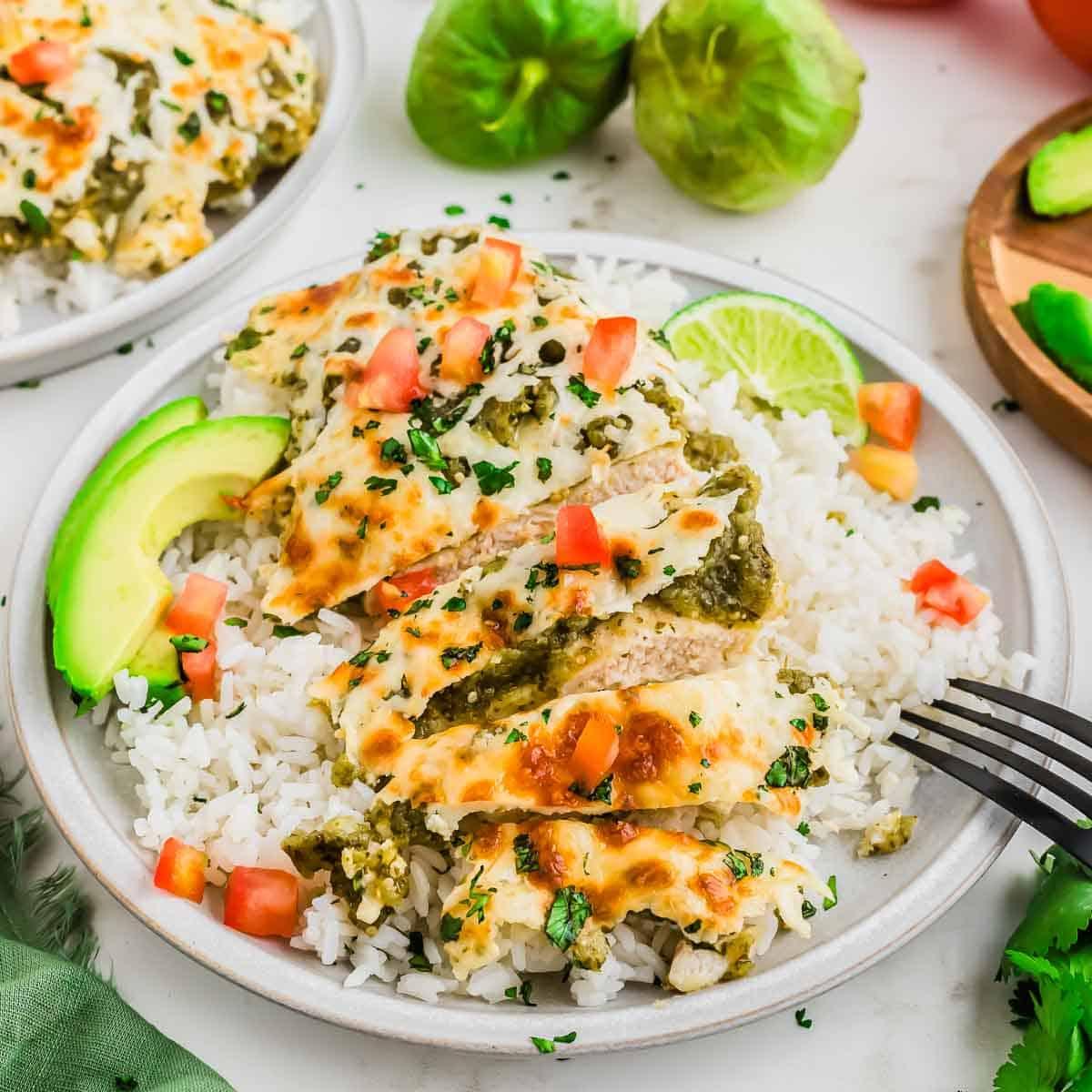 A plate of white rice topped with sliced chicken breast, melted cheese, salsa verde, diced tomatoes, chopped cilantro, and avocado, garnished with a lime wedge. A fork rests on the plate; tomatillos and avocado are in the background.