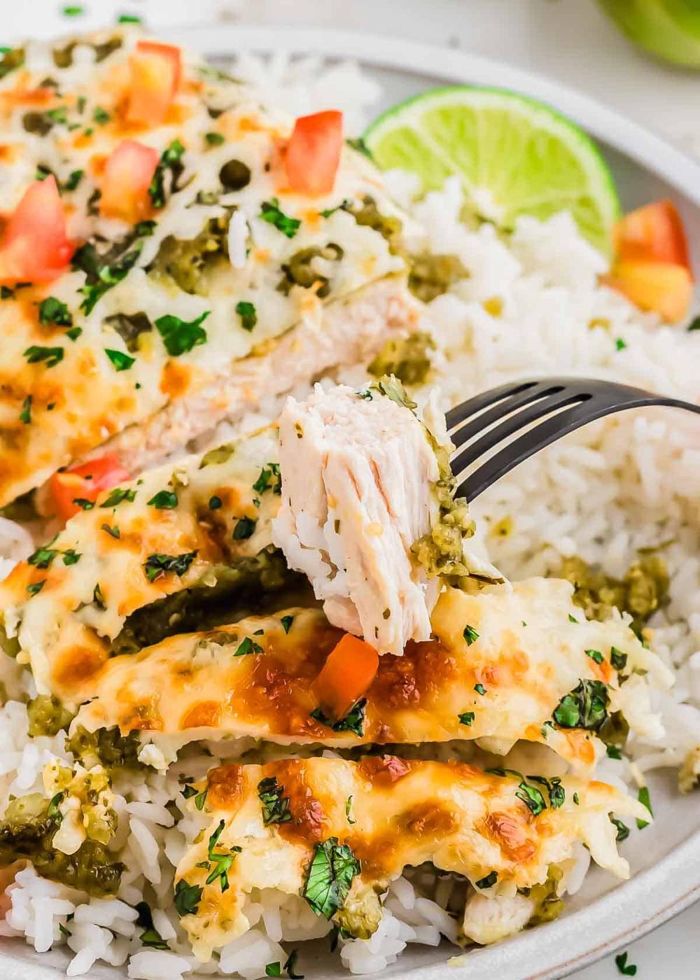 A close-up of a plate with sliced baked chicken covered in green salsa and melted cheese, served over white rice, garnished with chopped tomatoes and cilantro. A fork holds a bite-sized piece of chicken. Lime slices are on the side.