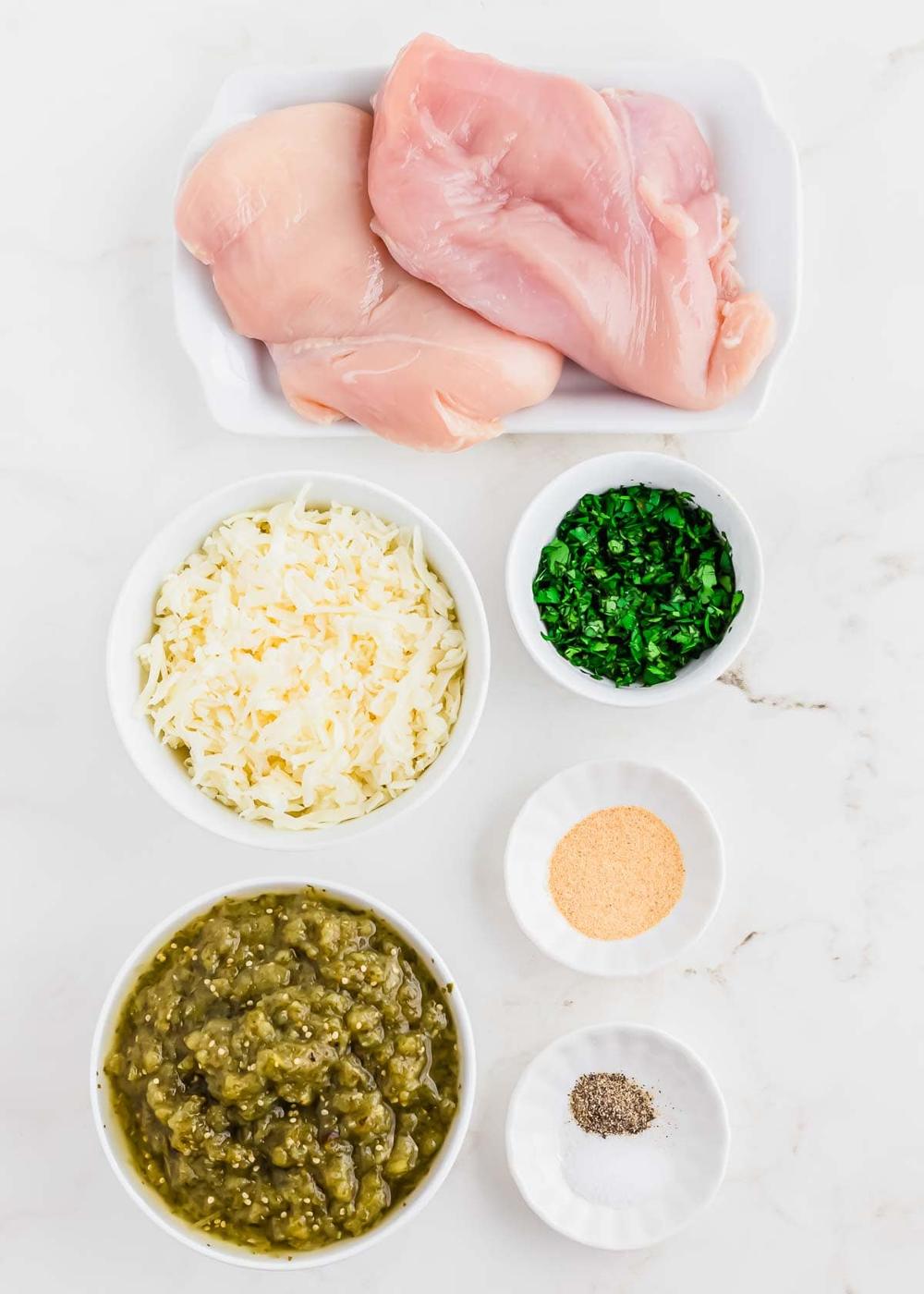Overhead view of raw chicken breasts, shredded cheese, chopped cilantro, salsa verde, garlic powder, and black pepper, each in separate bowls on a white surface.