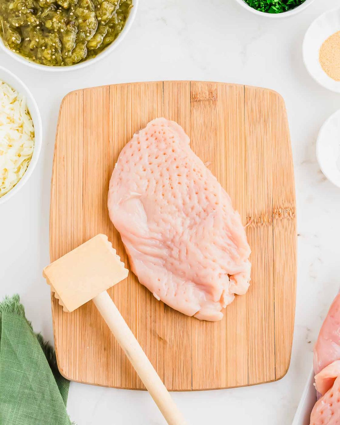 A raw chicken breast on a wooden cutting board, tenderized with a meat mallet, surrounded by bowls of ingredients such as green salsa, shredded cheese, and spices on a white countertop.