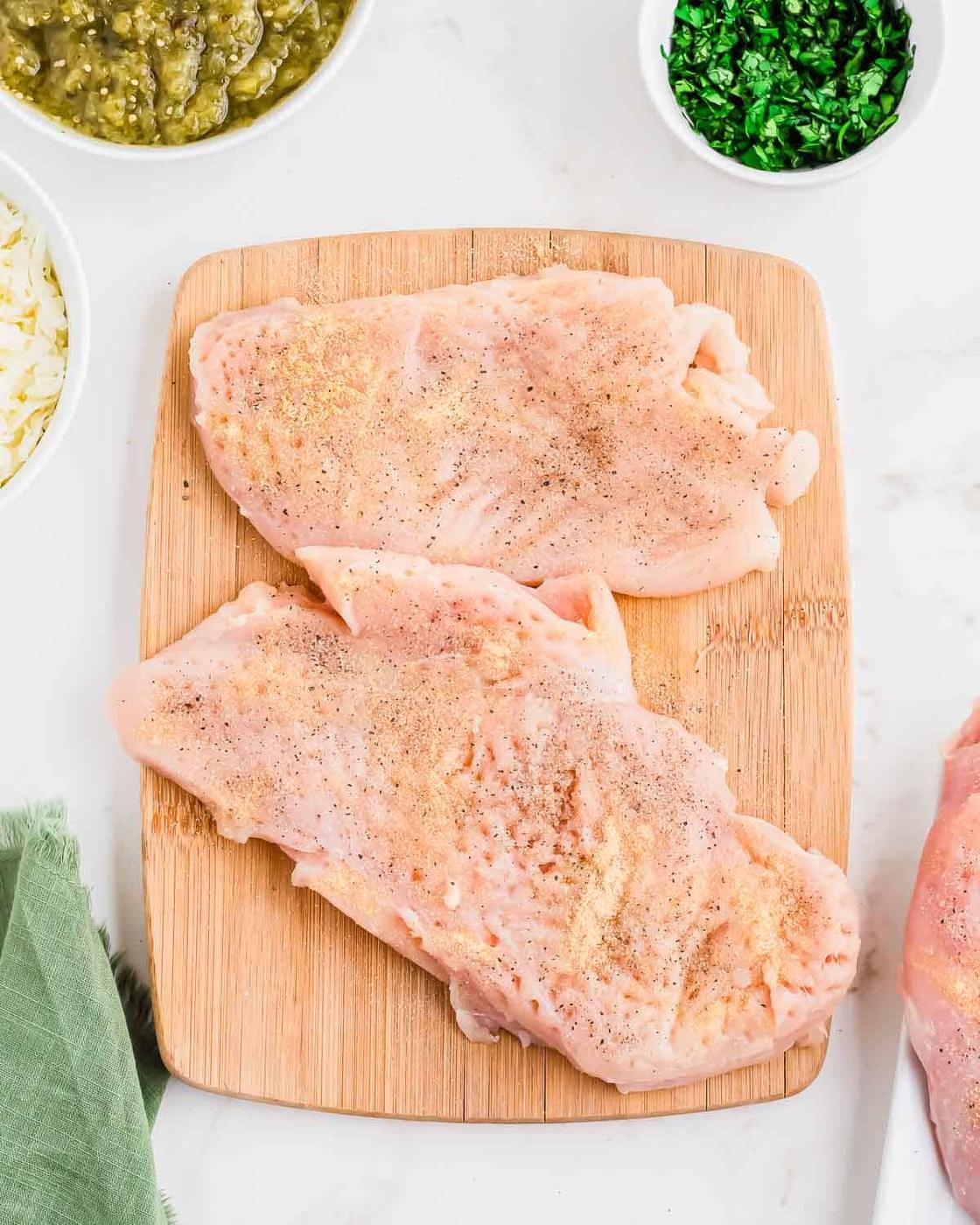 Two raw, seasoned chicken breasts on a wooden cutting board, surrounded by small bowls of green salsa, chopped herbs, and shredded cheese on a white surface.