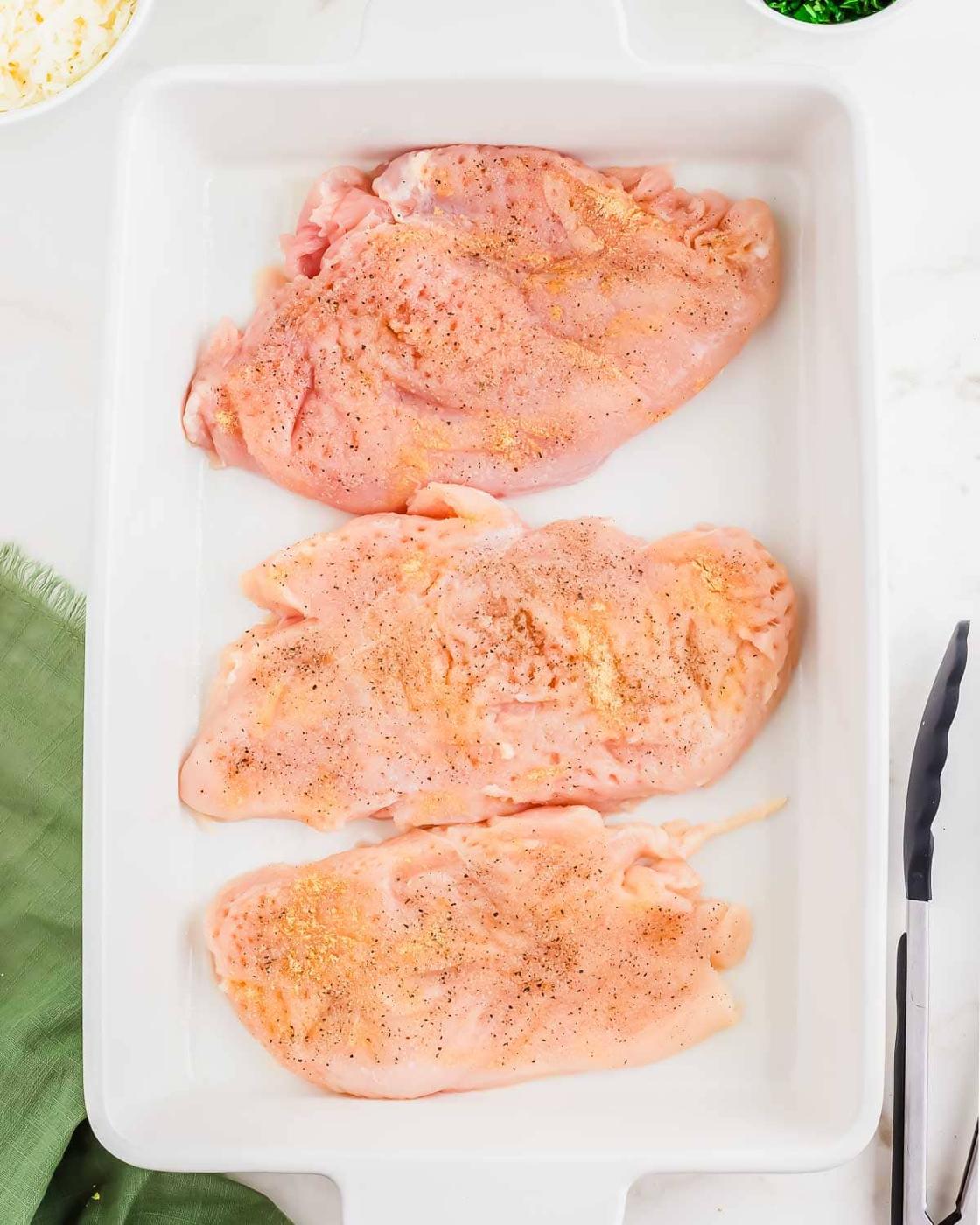 Three raw, seasoned chicken breasts arranged in a white baking dish, with salt, pepper, and spices visible on top. A green cloth, tongs, and ingredients are partially visible around the dish.