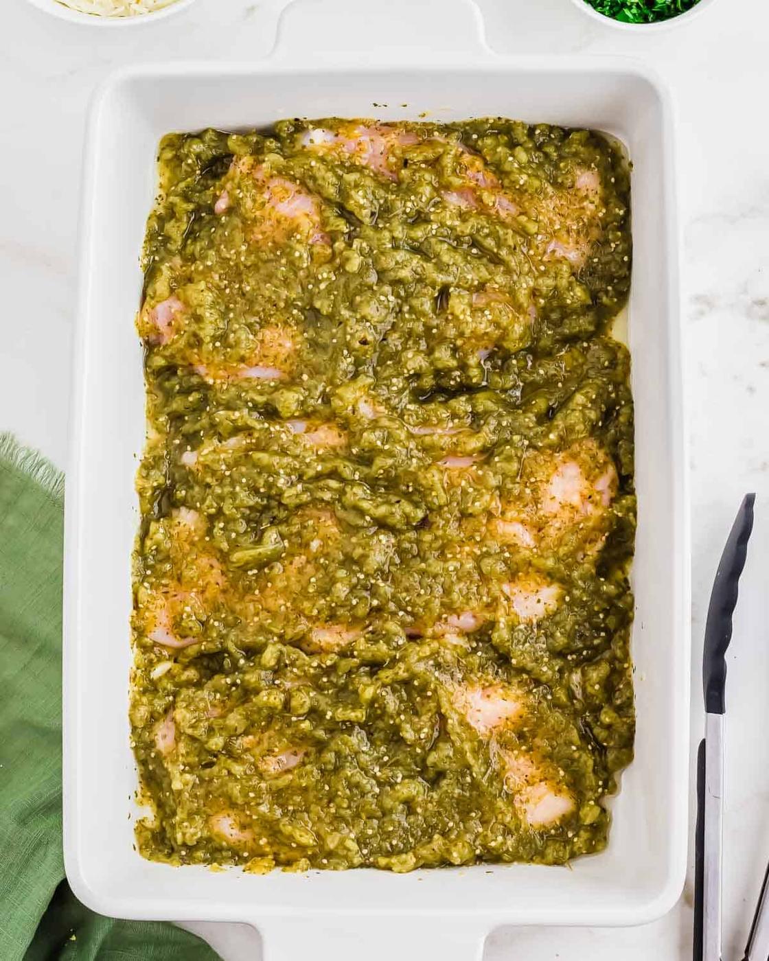 Raw chicken breasts in a white rectangular baking dish, covered with a chunky green salsa verde sauce, ready to be cooked. A green napkin and metal tongs are nearby on a white countertop.