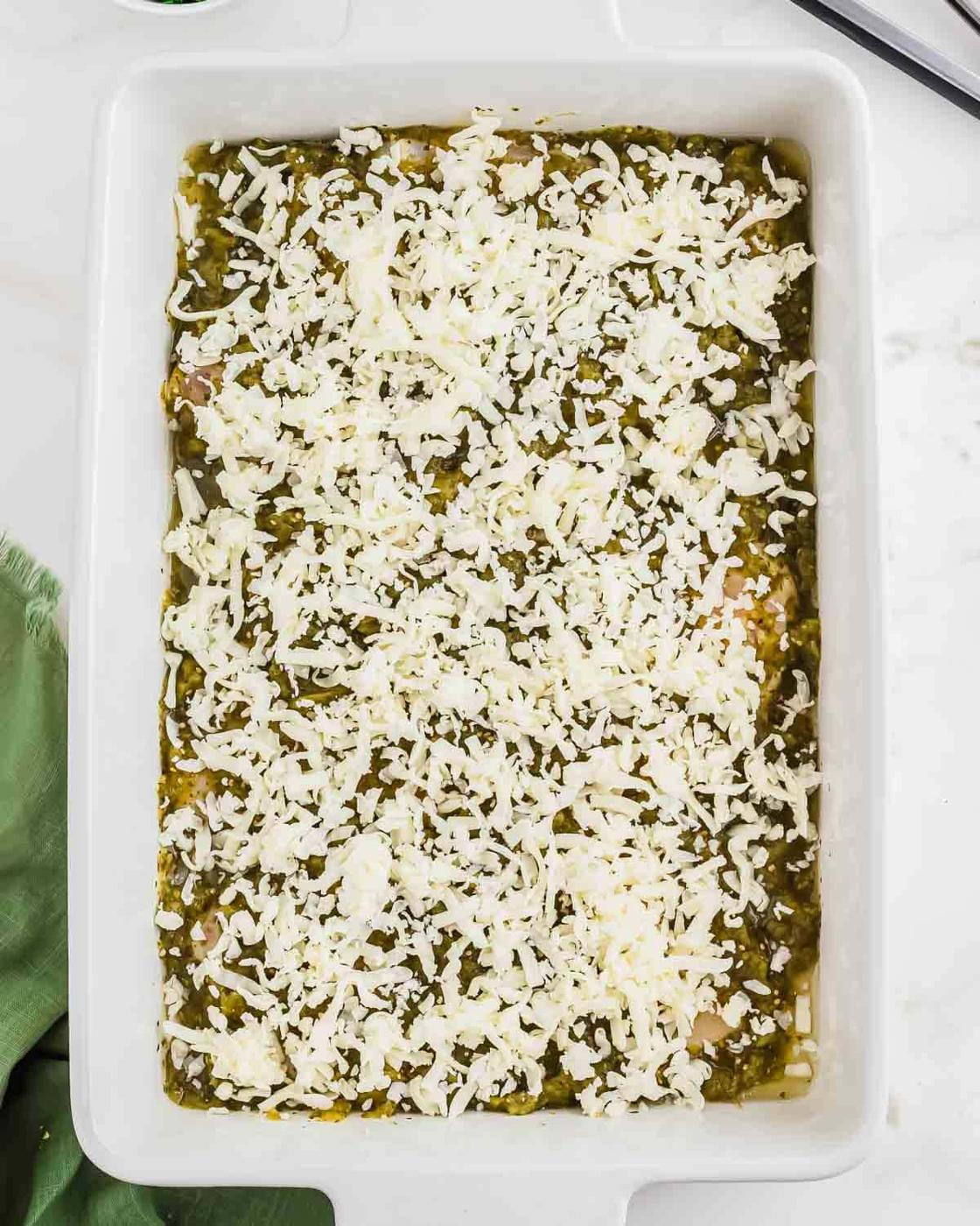 A white baking dish filled with a layered casserole, topped with shredded cheese and a green salsa, ready to be baked.