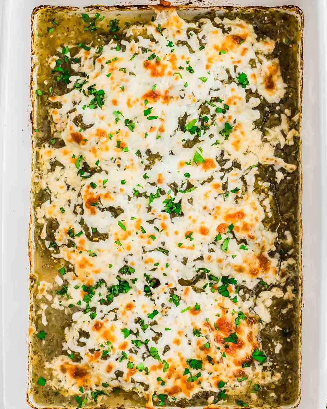 A baked casserole dish filled with salsa verde chicken topped with melted, golden-brown cheese and garnished with chopped fresh herbs.
