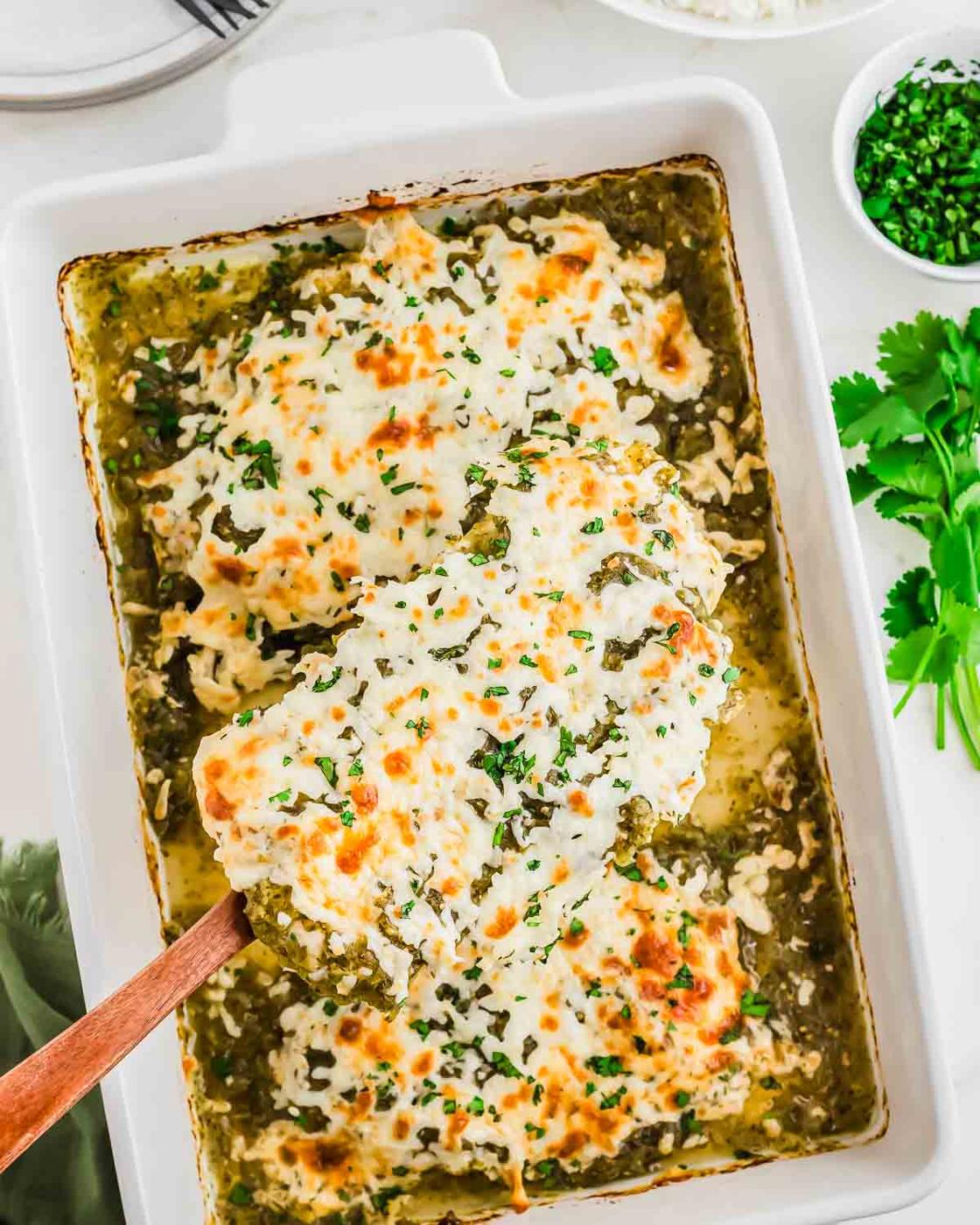 A baking dish filled with cheesy salsa verde chicken topped with melted cheese and chopped herbs, with a wooden spatula lifting a serving. Fresh cilantro and a bowl of chopped herbs are nearby.