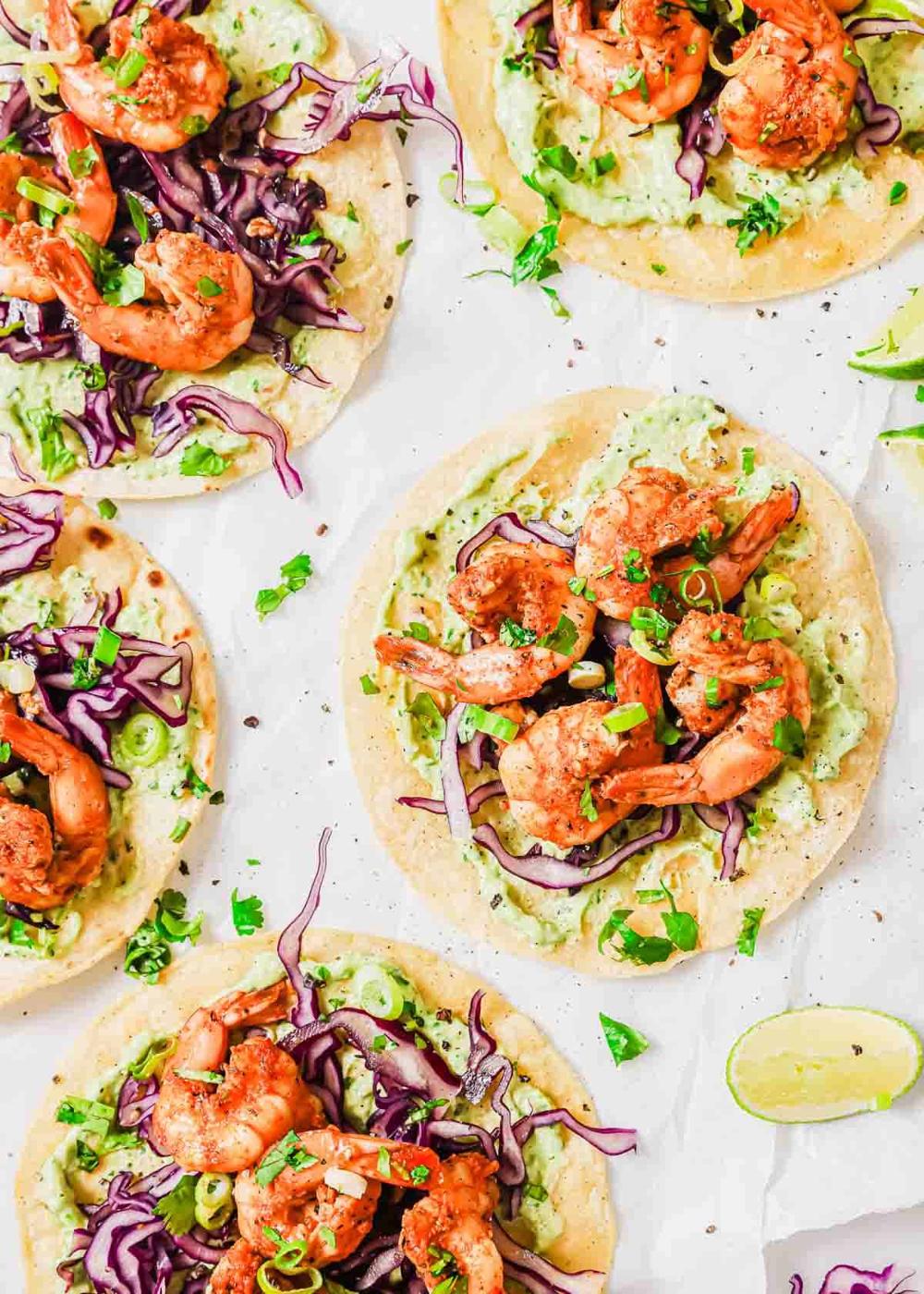 Five shrimp tostadas topped with shredded red cabbage, green onions, and cilantro on a white surface, with a lime wedge on the side.
