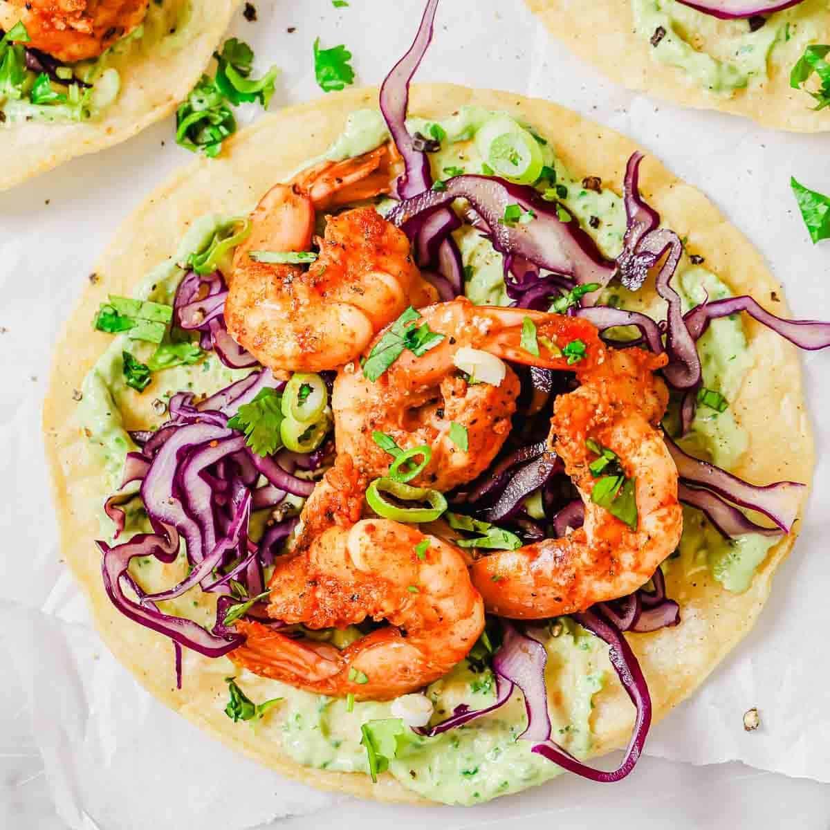 A tostada with seasoned shrimp, shredded red cabbage, green onions, cilantro, and guacamole on a tortilla.