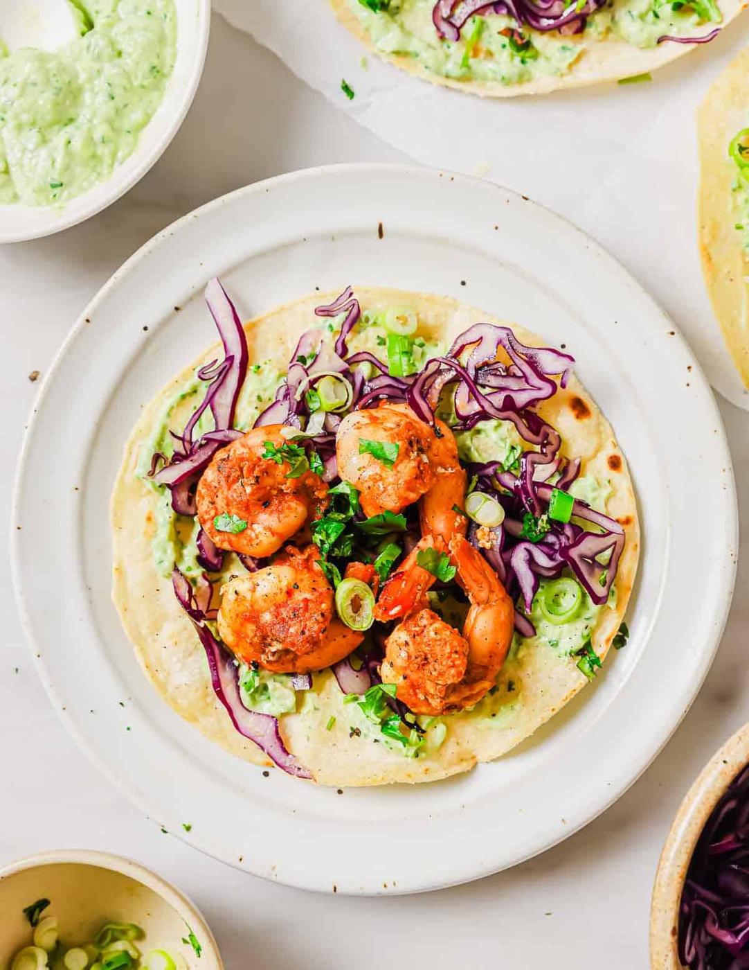 A plate with a tostada topped with grilled shrimp, shredded red cabbage, chopped green onions, and cilantro. Surrounding the plate are additional taco ingredients and dishes.