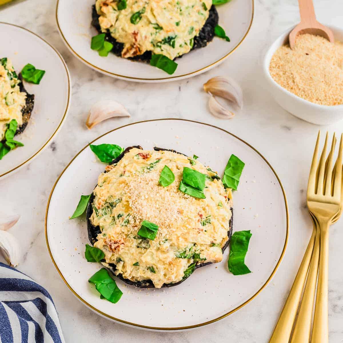 A plate featuring a stuffed portobello mushroom cap topped with a creamy mixture and sprinkled with breadcrumbs. Fresh basil leaves are scattered on top, and there's a small bowl of breadcrumbs and garlic cloves nearby on a marble surface. Golden cutlery is beside the plate.