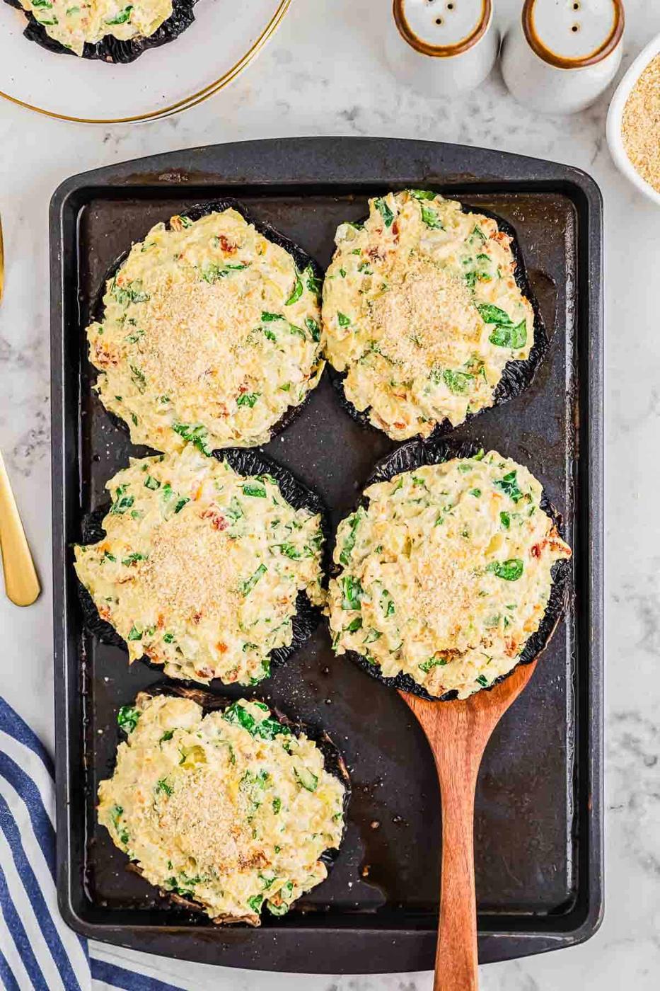 Five stuffed portobello mushrooms topped with breadcrumbs on a baking tray. One is being lifted with a wooden spatula. The filling appears creamy with bits of spinach and seasonings. A striped napkin and salt shakers are in the background.