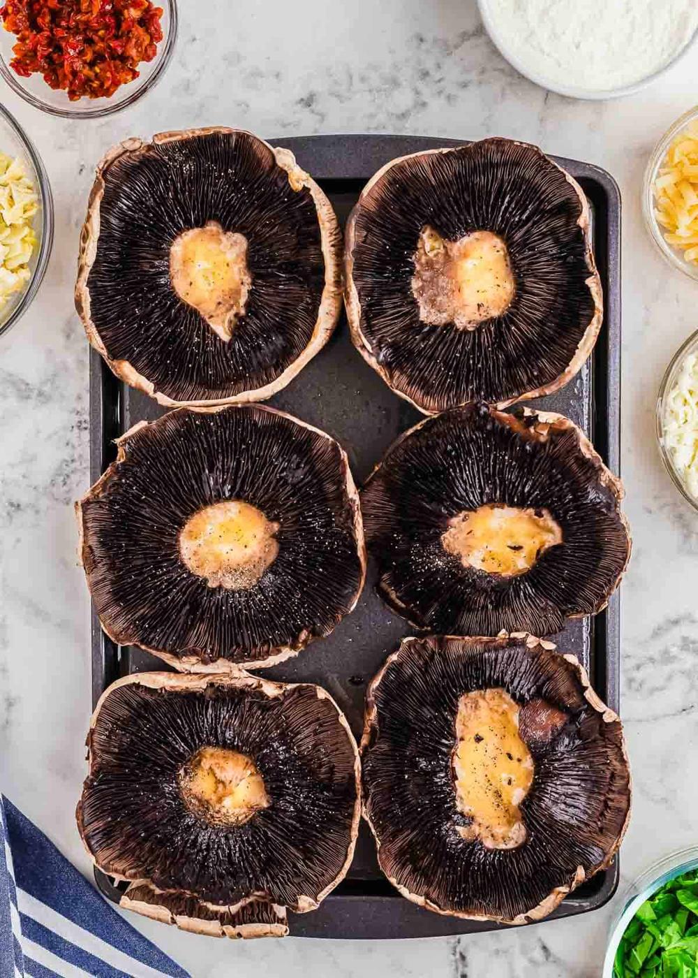 Six large portobello mushrooms with gills exposed are placed on a baking tray. Ingredients like chopped tomatoes, cheese, and chopped green onions are visible around the tray, suggesting preparation for a dish.