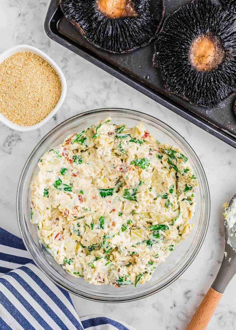 A glass bowl filled with creamy spinach and artichoke filling is set on a marble surface. Next to it is a small bowl of breadcrumbs and a baking tray with large portobello mushroom caps. A blue and white striped cloth is nearby.