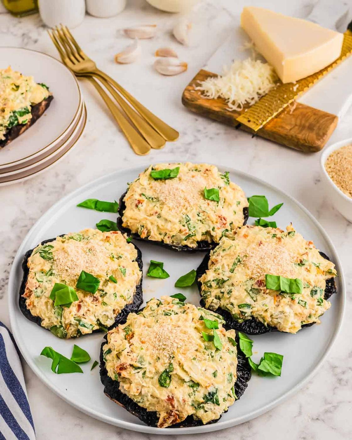 A plate with four creamy stuffed portobello mushrooms garnished with fresh basil leaves. The table also has plates, forks, garlic cloves, grated cheese, a block of cheese, and breadcrumbs in the background.