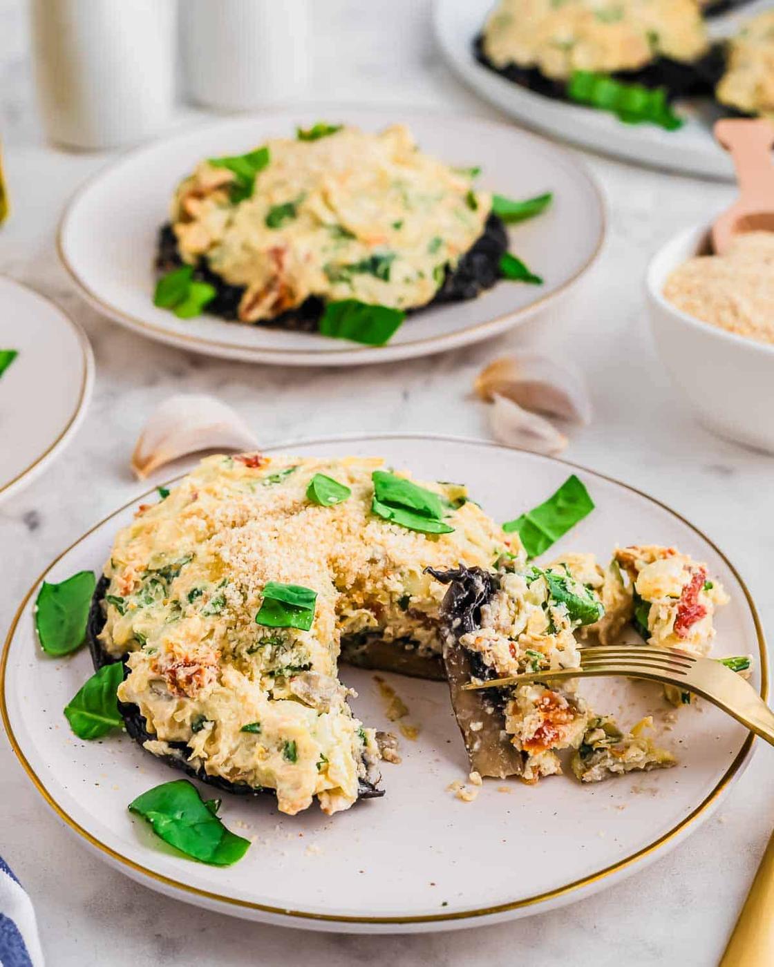 A plate with a creamy spinach and artichoke stuffed mushroom, topped with grated cheese and fresh basil leaves. A fork rests on the plate, and there are more similar plates and ingredients visible in the background.