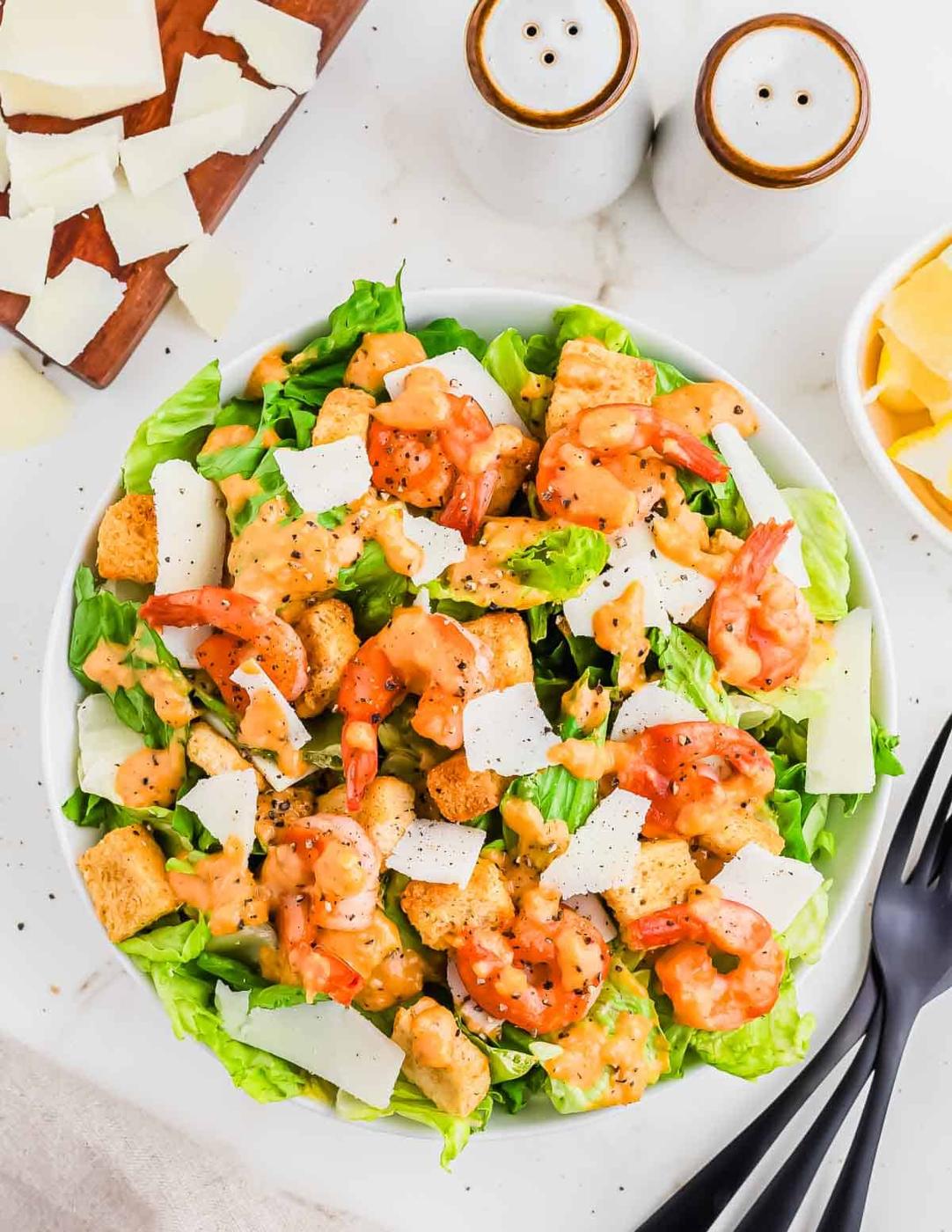 A bowl of Caesar salad topped with shrimp, croutons, shaved parmesan, and dressing, with utensils, salt and pepper shakers, and extra cheese slices nearby.