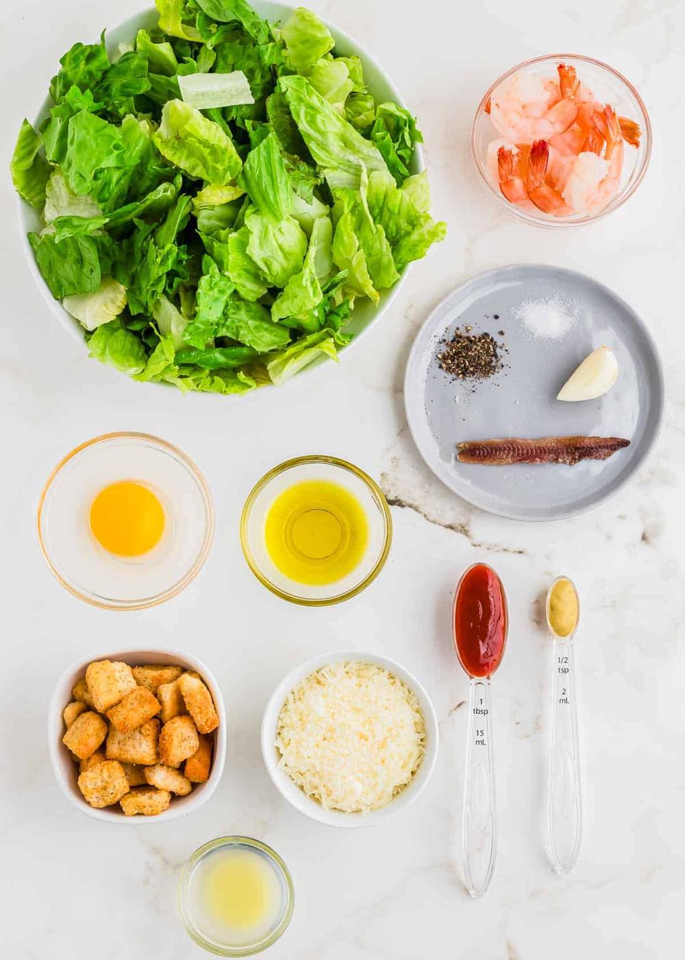 Ingredients for Caesar salad including a bowl of romaine lettuce, shrimp, croutons, grated cheese, anchovies, garlic, mustard, mayonnaise, lemon juice, oil, a raw egg, salt, and pepper.