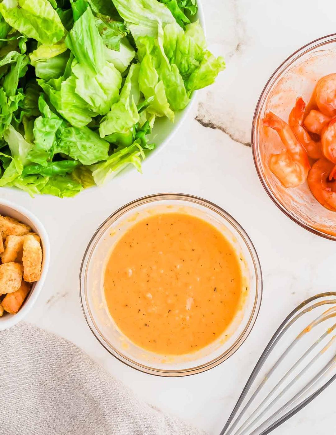 A bowl of lettuce, peeled shrimp in another bowl, a small bowl with croutons, a bowl of sriracha Caesar dressing, and a whisk on a white marble surface.