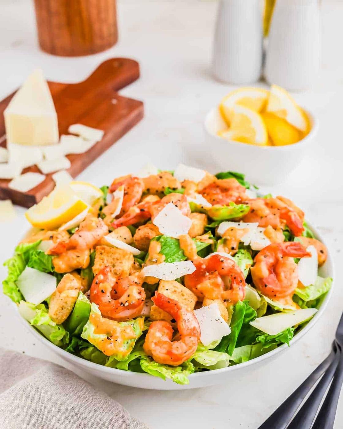 A bowl of shrimp Caesar salad with lettuce, grilled shrimp, shaved Parmesan cheese, and lemon wedges. A cutting board with Parmesan cheese and another bowl of lemon wedges are in the background.