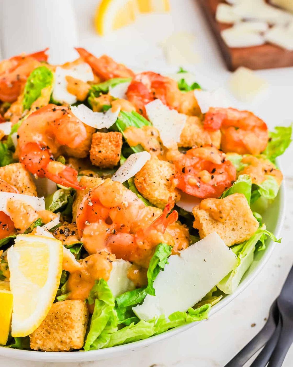 A close-up of a shrimp Caesar salad with croutons, romaine lettuce, shaved Parmesan cheese, and lemon wedges in a white bowl.
