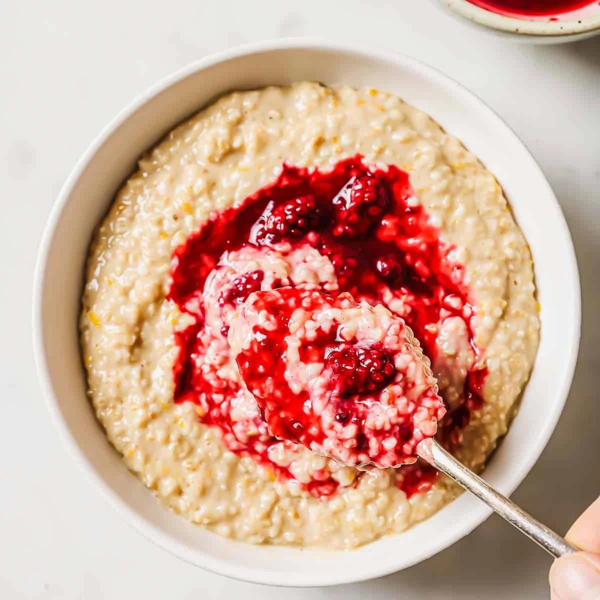 A bowl of steel cut oatmeal topped with a vibrant blackberry orange compote. A spoon with oatmeal and berry compote is resting inside the bowl, showing the thick texture and rich red color contrasting with the creamy oatmeal.