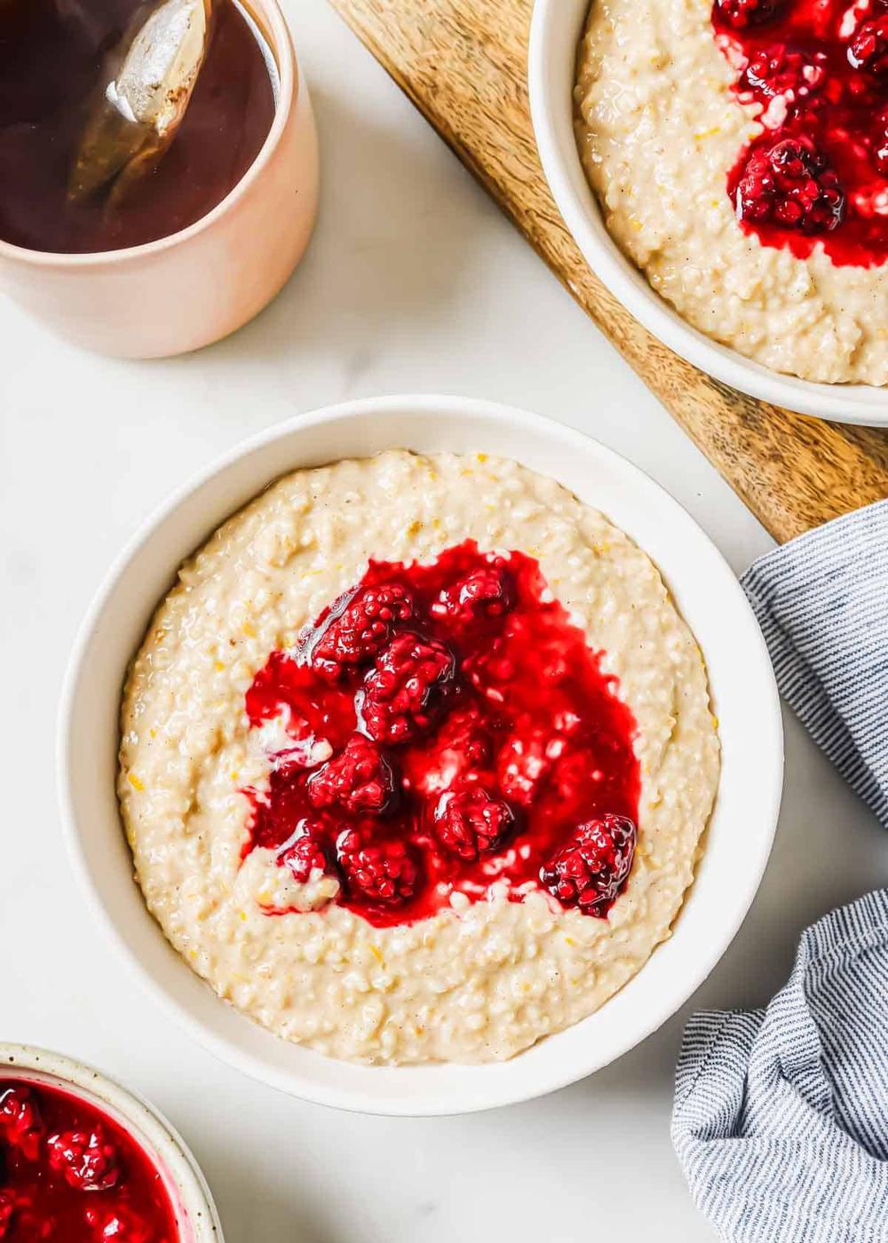 A bowl of steel cut oatmeal topped with blackberry compote, surrounded by another bowl of oatmeal, a cup of tea, and a striped cloth on a light surface. The compote appears rich and glossy, adding a pop of color to the creamy oatmeal.