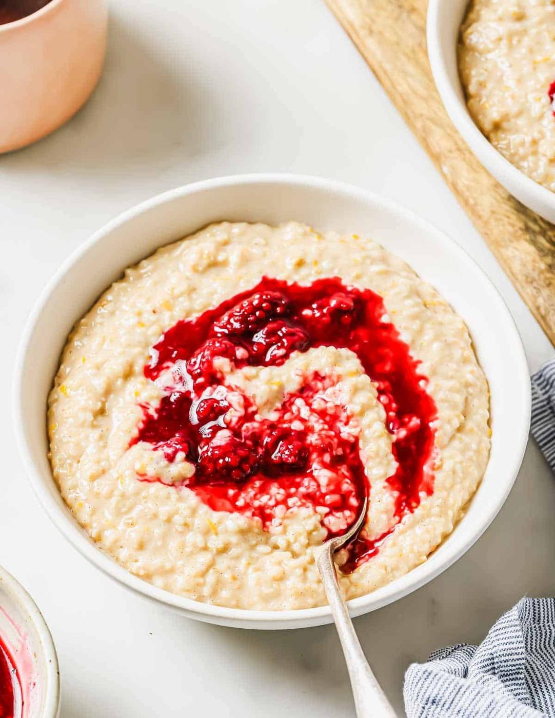 A bowl of creamy oatmeal topped with a swirl of vibrant berry compote. A spoon rests in the bowl, and there's a glimpse of a napkin beside it. The setting is on a light-colored surface.