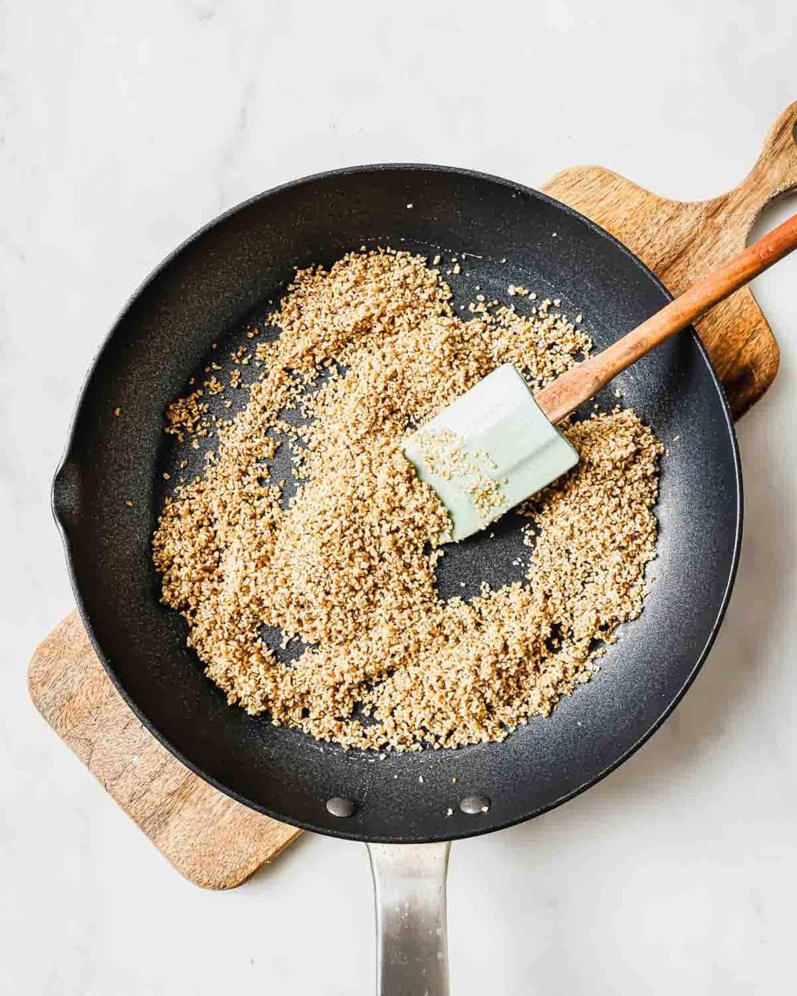 A black frying pan with toasted steel cut oatmeal being stirred by a spatula. The pan is on a wooden cutting board against a light gray background.