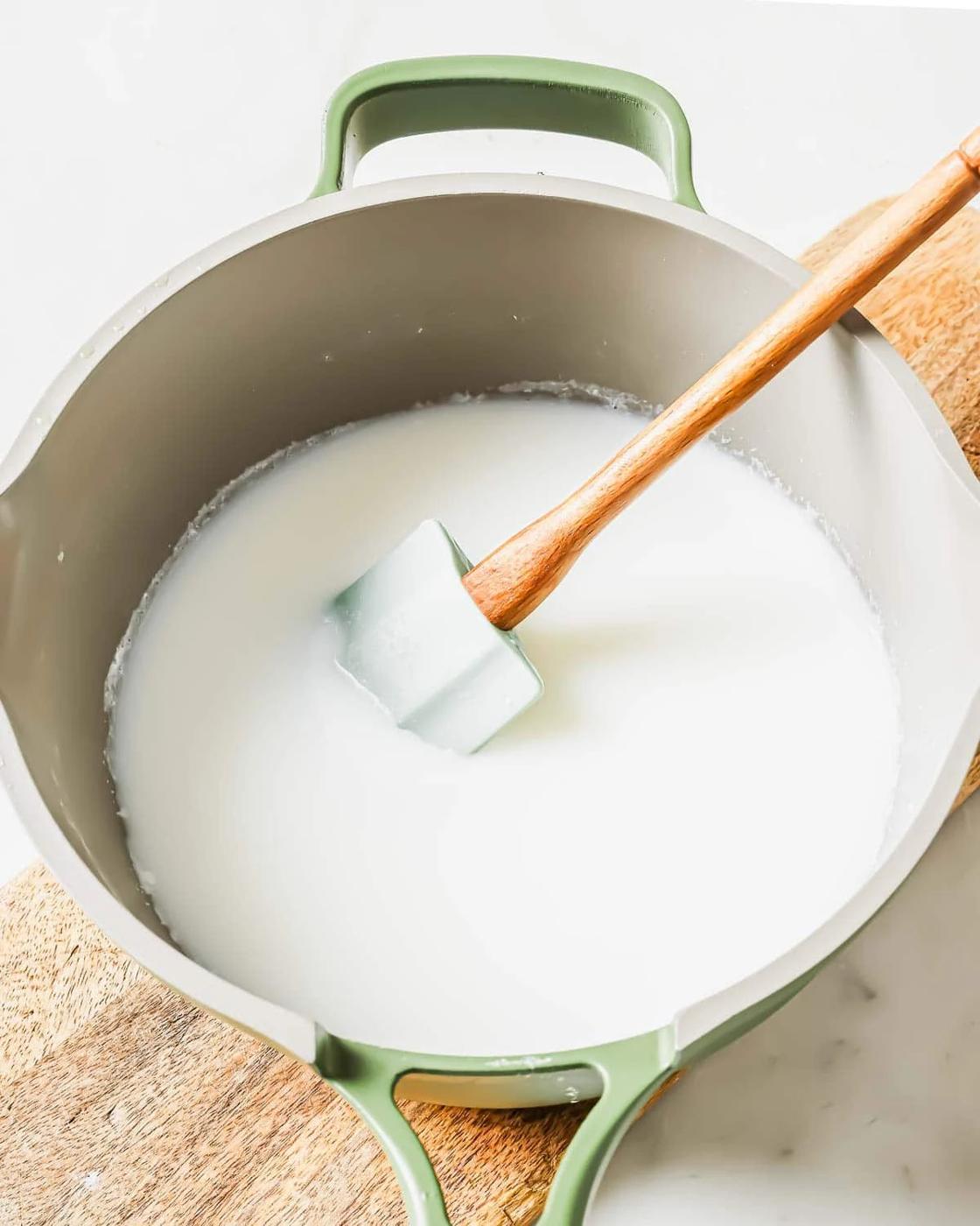 A green pot containing a milky white liquid sits on a wooden surface. A wooden spatula with a light green silicone tip is placed inside the pot, leaning against the edge. The scene suggests cooking or mixing ingredients.