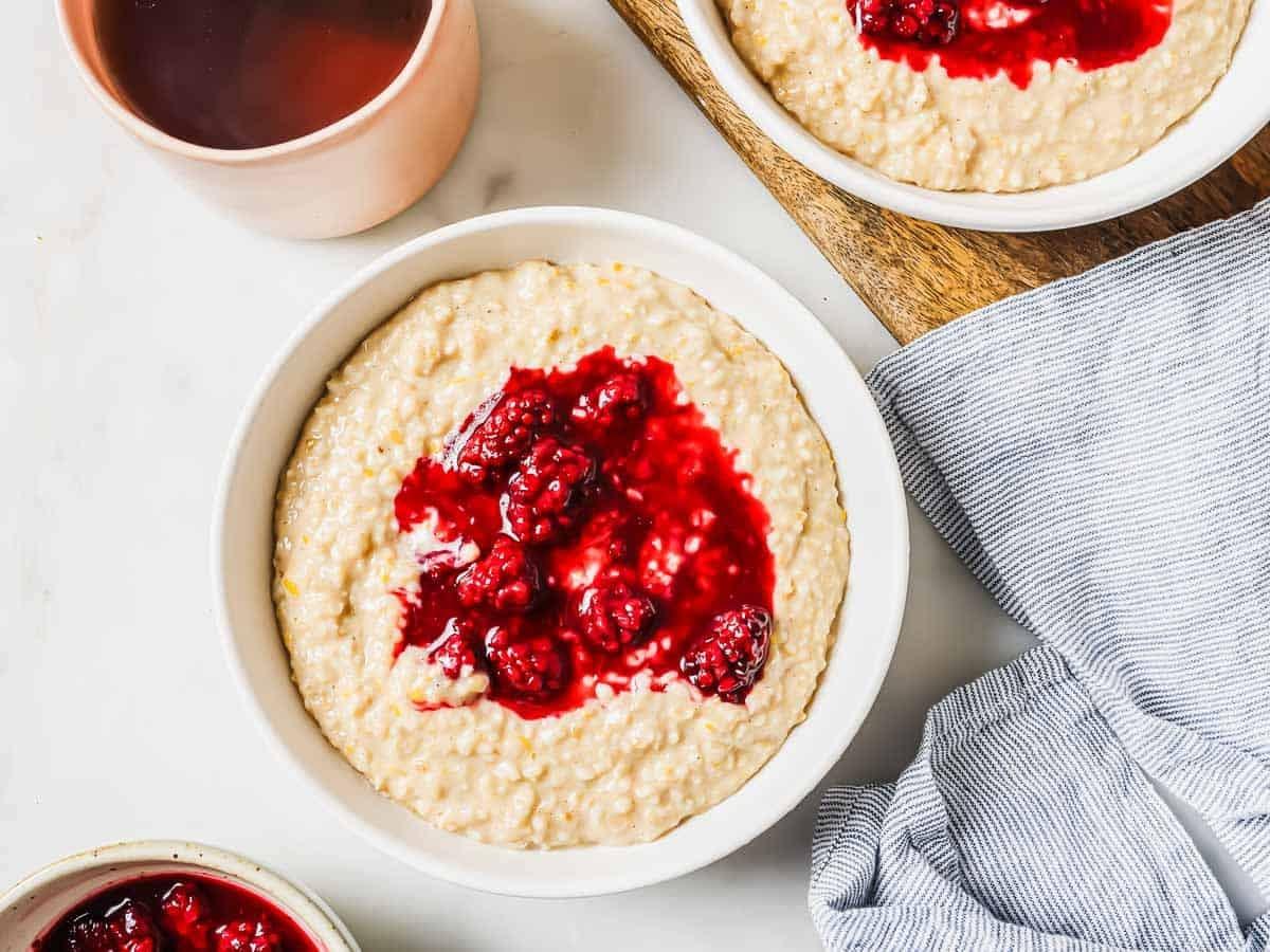 A bowl of creamy hearty steel cut oatmeal topped with a vibrant blackberry sauce on a white table. A second bowl of oatmeal and a cup of tea are nearby, along with a striped cloth napkin. The scene has a cozy and inviting feel.