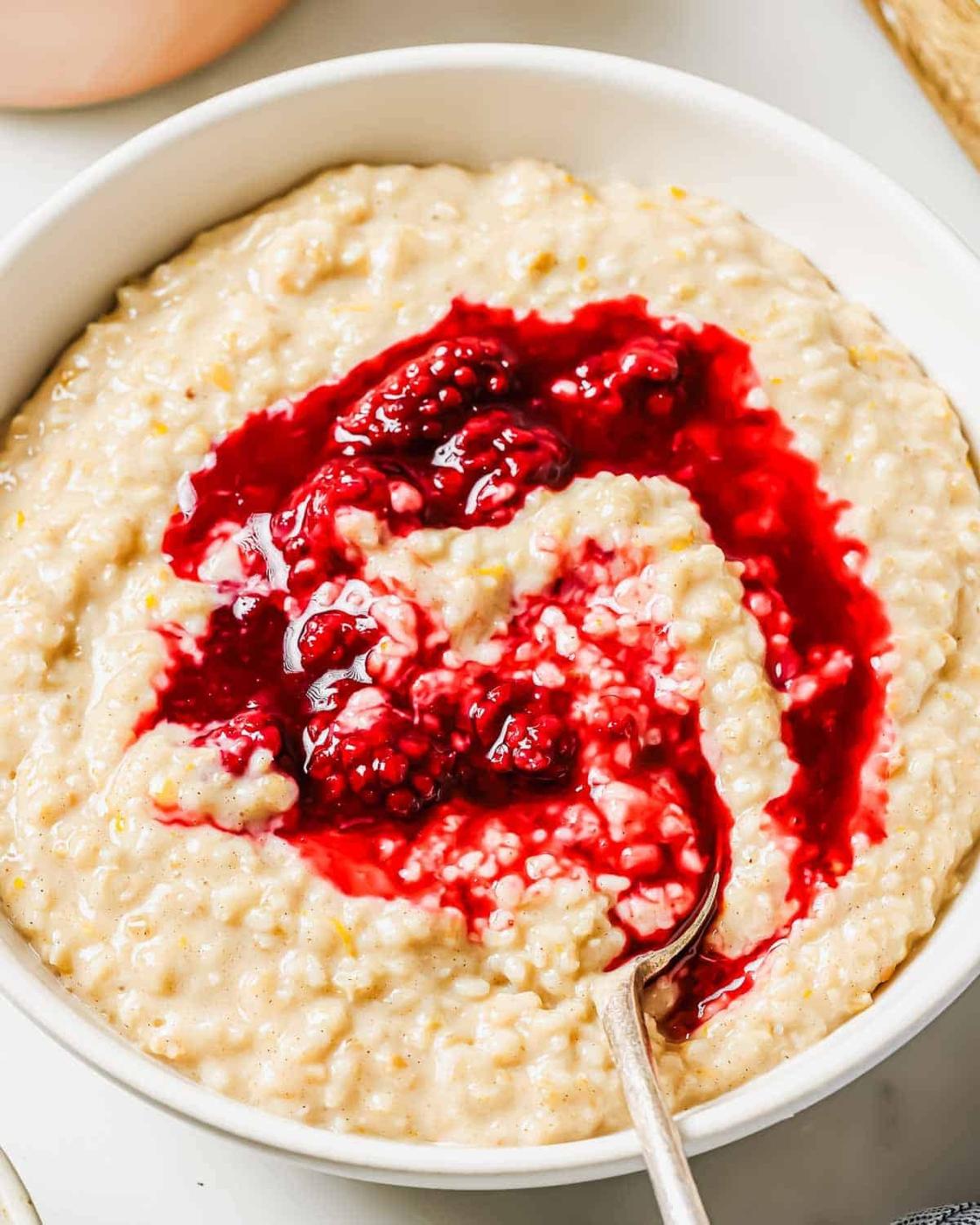 A bowl of steel cut oatmeal topped with a swirl of bright blackberry compote. The oatmeal appears creamy and thick, while the compote adds a vibrant contrast. A spoon rests in the bowl.