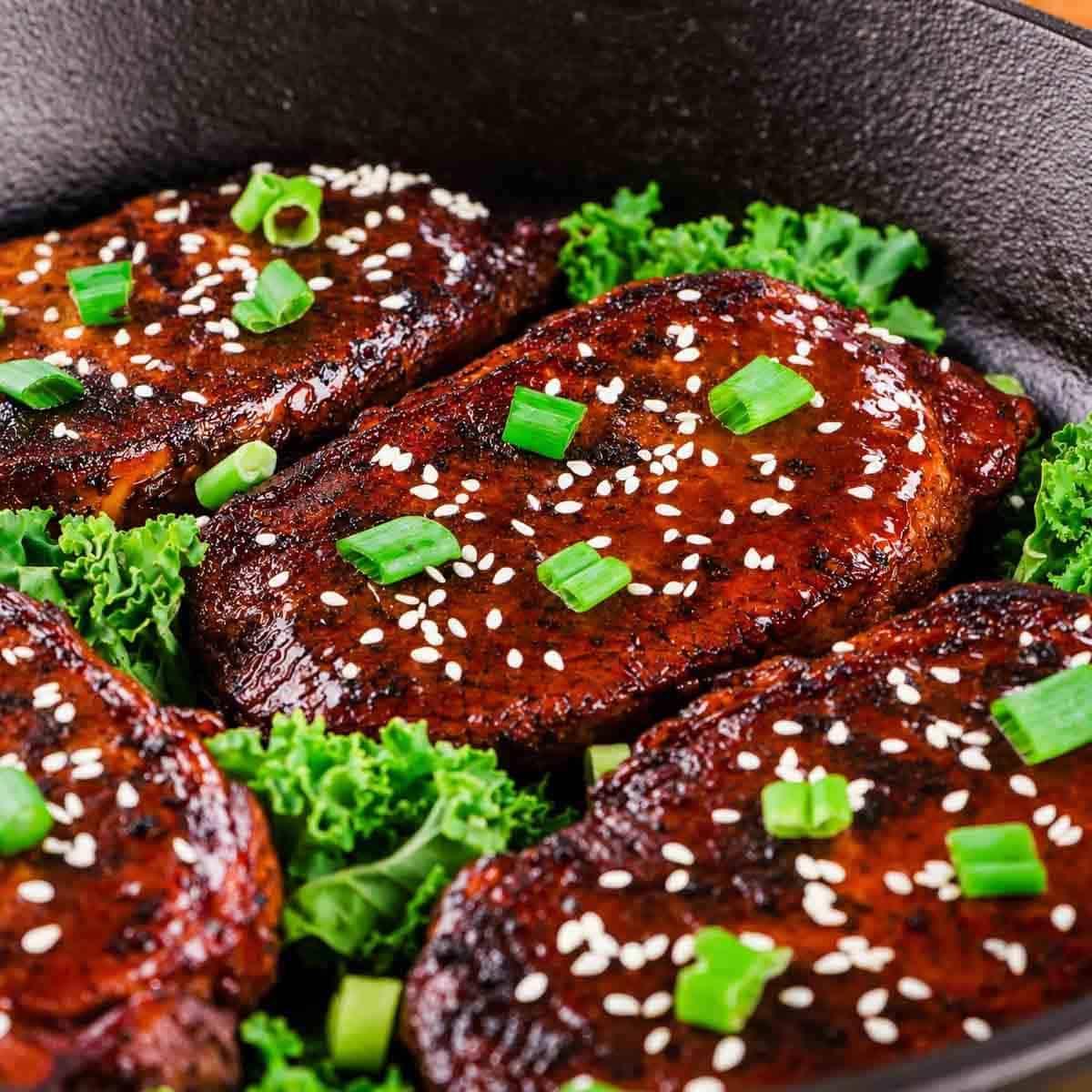 Four pieces of honey garlic glazed, seared pork topped with sesame seeds and chopped green onions sit on a bed of fresh green kale in a black skillet.