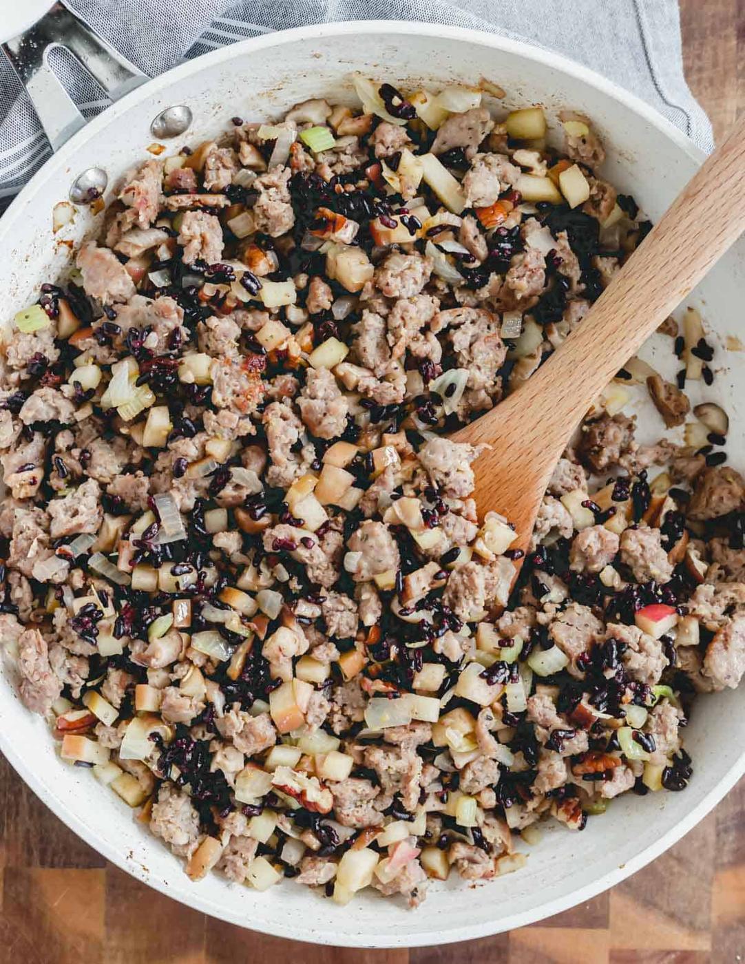 Sausage, mushroom, apple, onion, raisin and pecan stuffing in a white skillet with a wooden spatula.