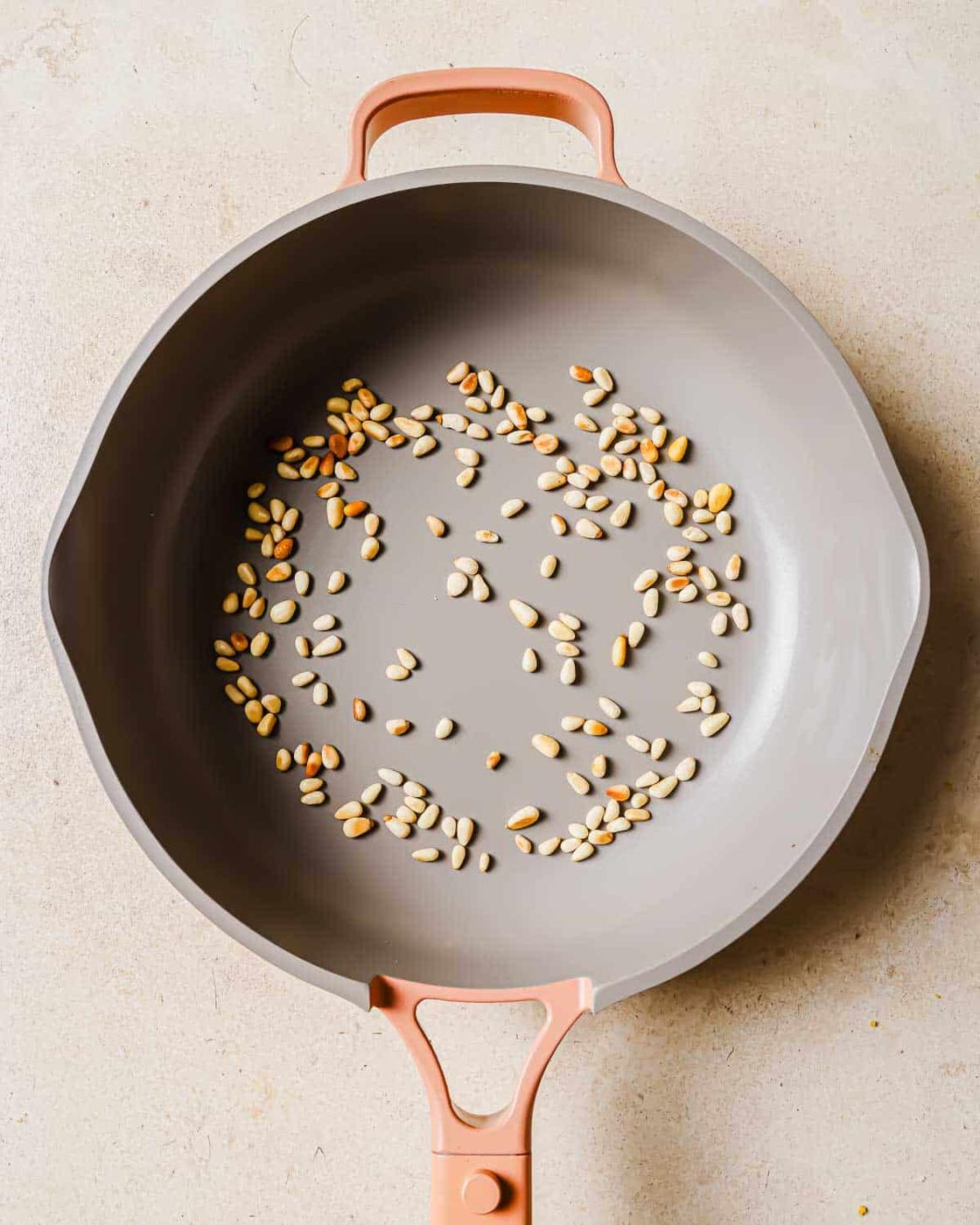 A few scattered pine nuts are toasting in a large gray frying pan with a pink handle, placed on a beige surface.