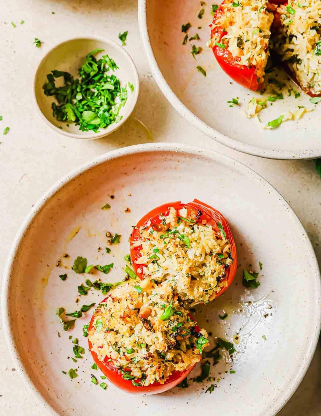 Two stuffed tomatoes filled with a mixture of rice and herbs are on a plate. A small bowl of finely chopped herbs is beside the plate.