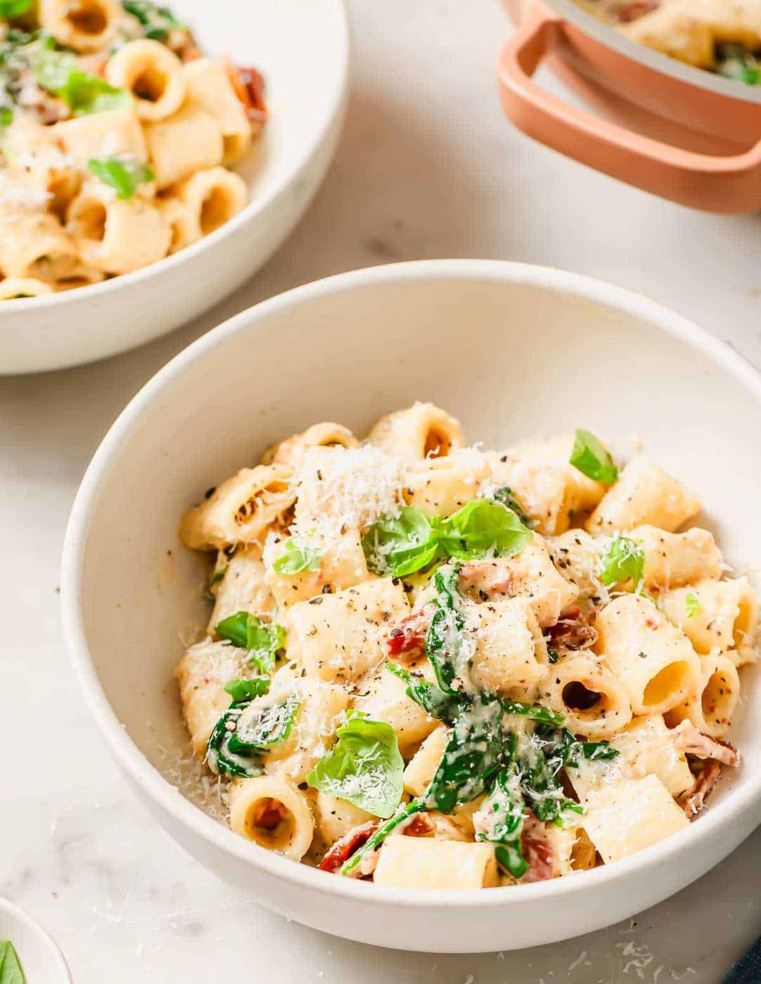 A bowl of pasta with cheese, basil, and sun-dried tomato, served in a white bowl.