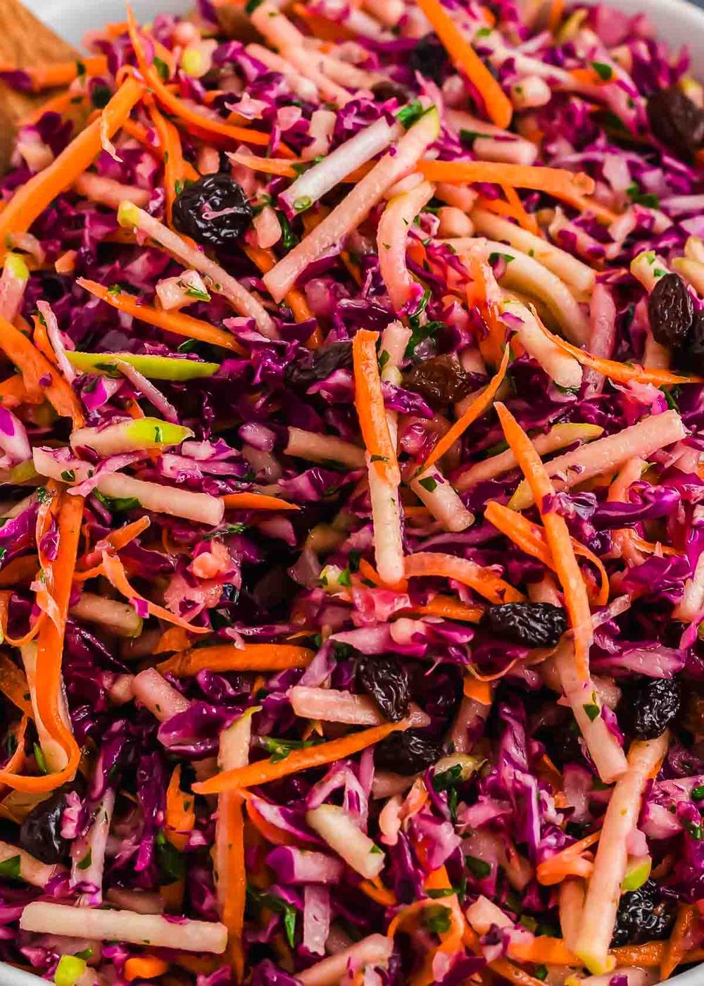 A close-up of a colorful slaw featuring shredded red cabbage, carrots, and apple, mixed with raisins and herbs. The vibrant salad has a fresh and crunchy appearance.