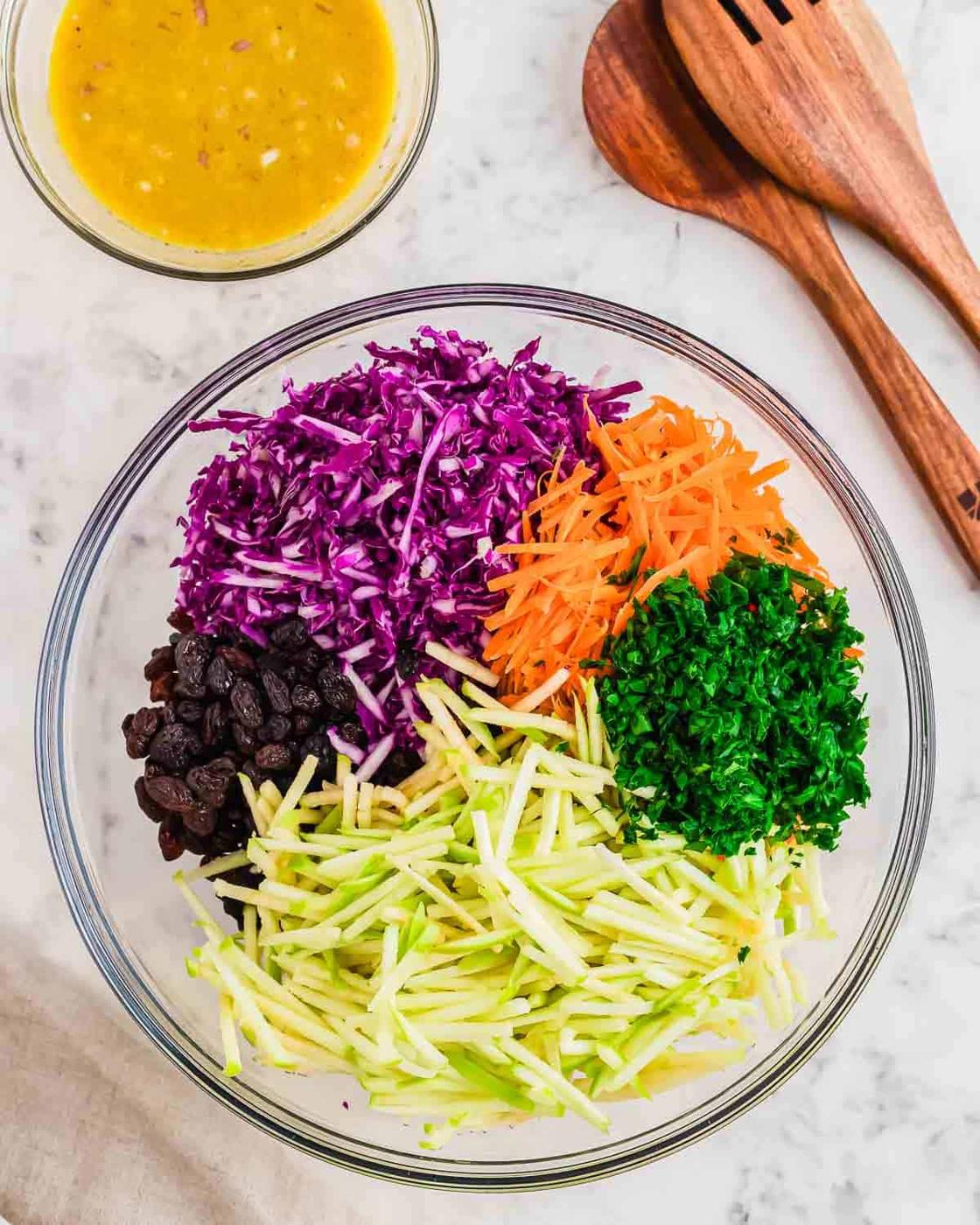 A glass bowl filled with red cabbage, grated carrots, raisins, chopped parsley, and shredded apples. Wooden salad servers are beside it on a marble surface. A small bowl of dressing is nearby.