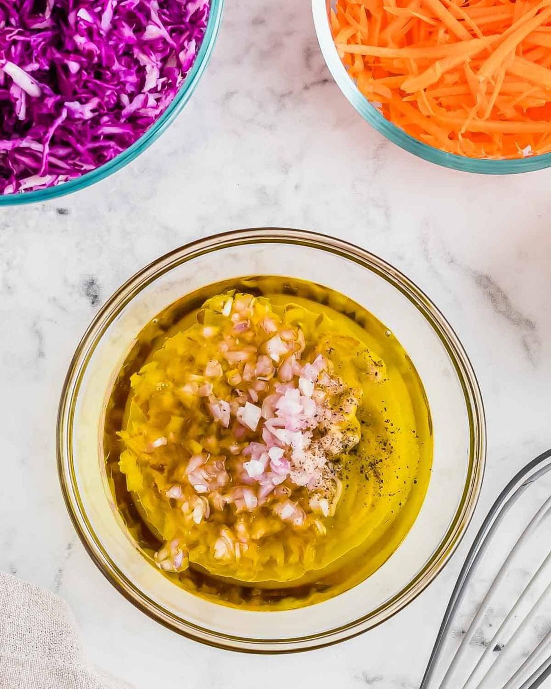A glass bowl filled with a mixture of olive oil, minced garlic, and finely chopped onions is placed on a marble surface. Nearby are bowls containing shredded purple cabbage and grated carrots. A whisk is partially visible in the lower corner.