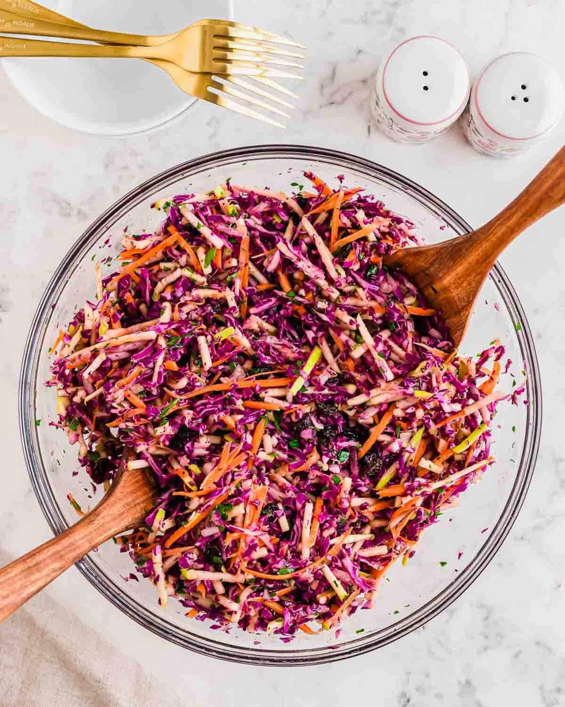 A bowl of colorful coleslaw with shredded red cabbage, carrots, and herbs, mixed with dressing. Wooden salad servers are placed in the bowl. Gold forks and salt and pepper shakers are on the marble countertop nearby.