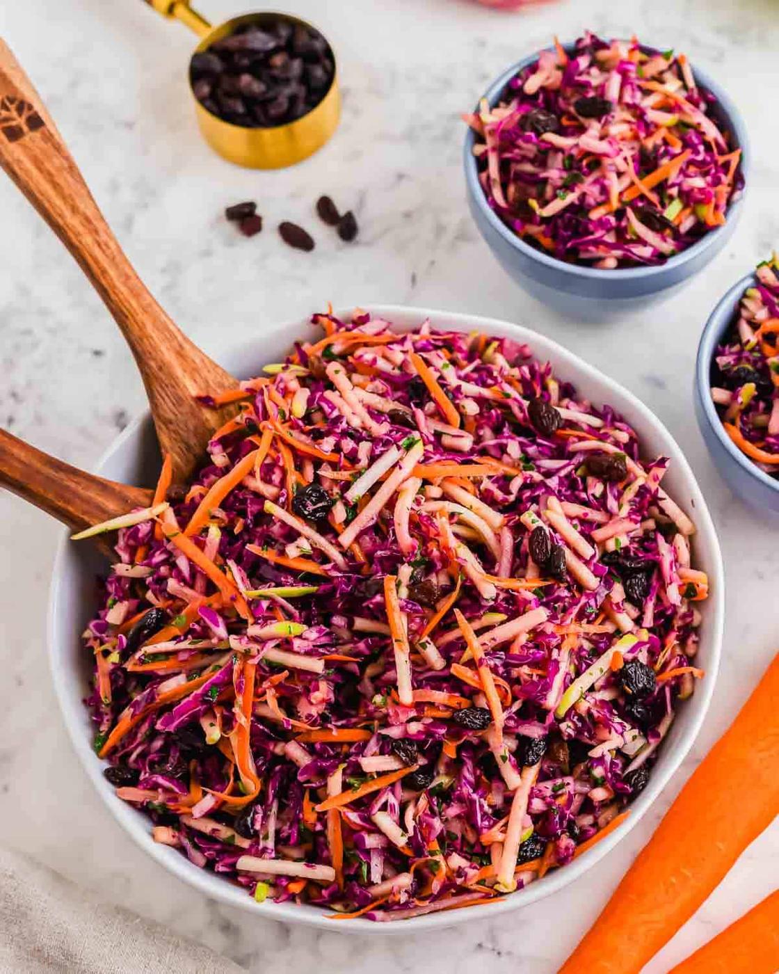 A bowl of vibrant coleslaw featuring shredded red cabbage, carrots, and raisins, placed on a marble surface. Two wooden serving spoons rest in the bowl. A smaller bowl and a cup of raisins are in the background.