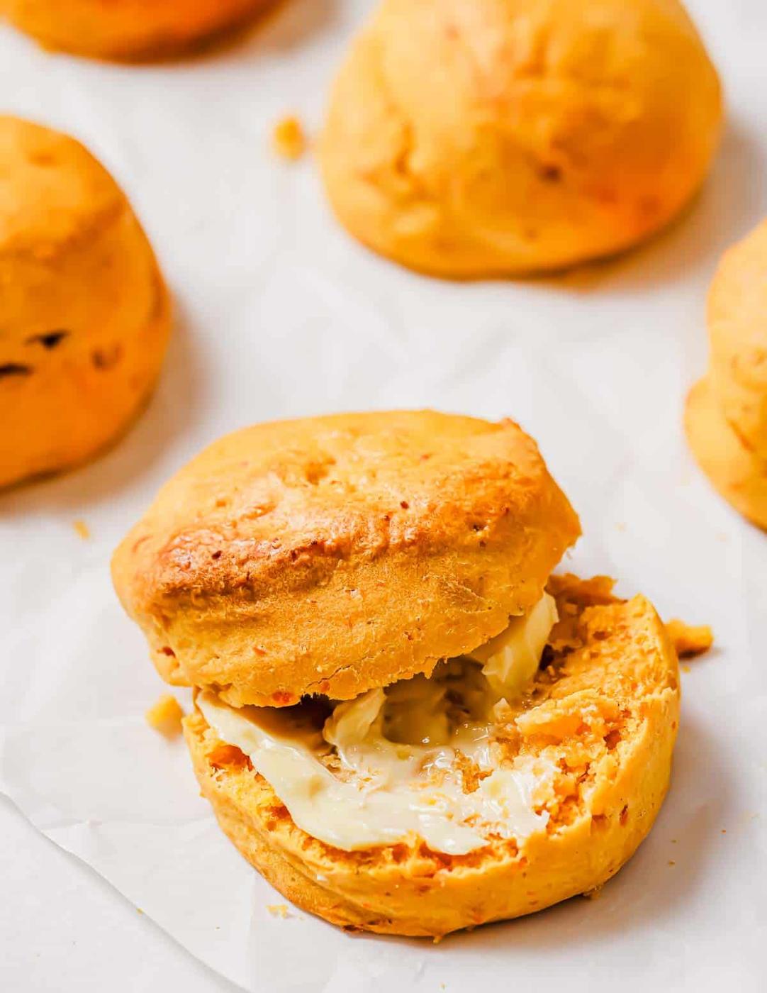 A close-up of a sweet potato biscuit split open with butter spread on the bottom half. Other biscuits are partially visible in the background on a sheet of parchment paper.