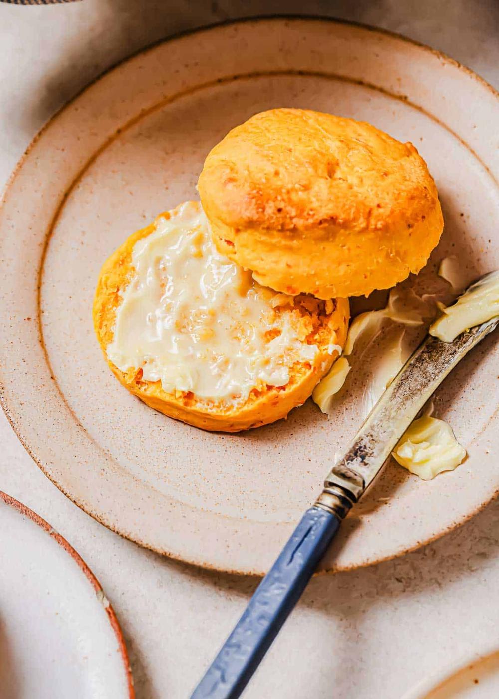 A sweet potato biscuit sliced in half with butter spread on the lower half, placed on a beige plate with a butter knife next to it.