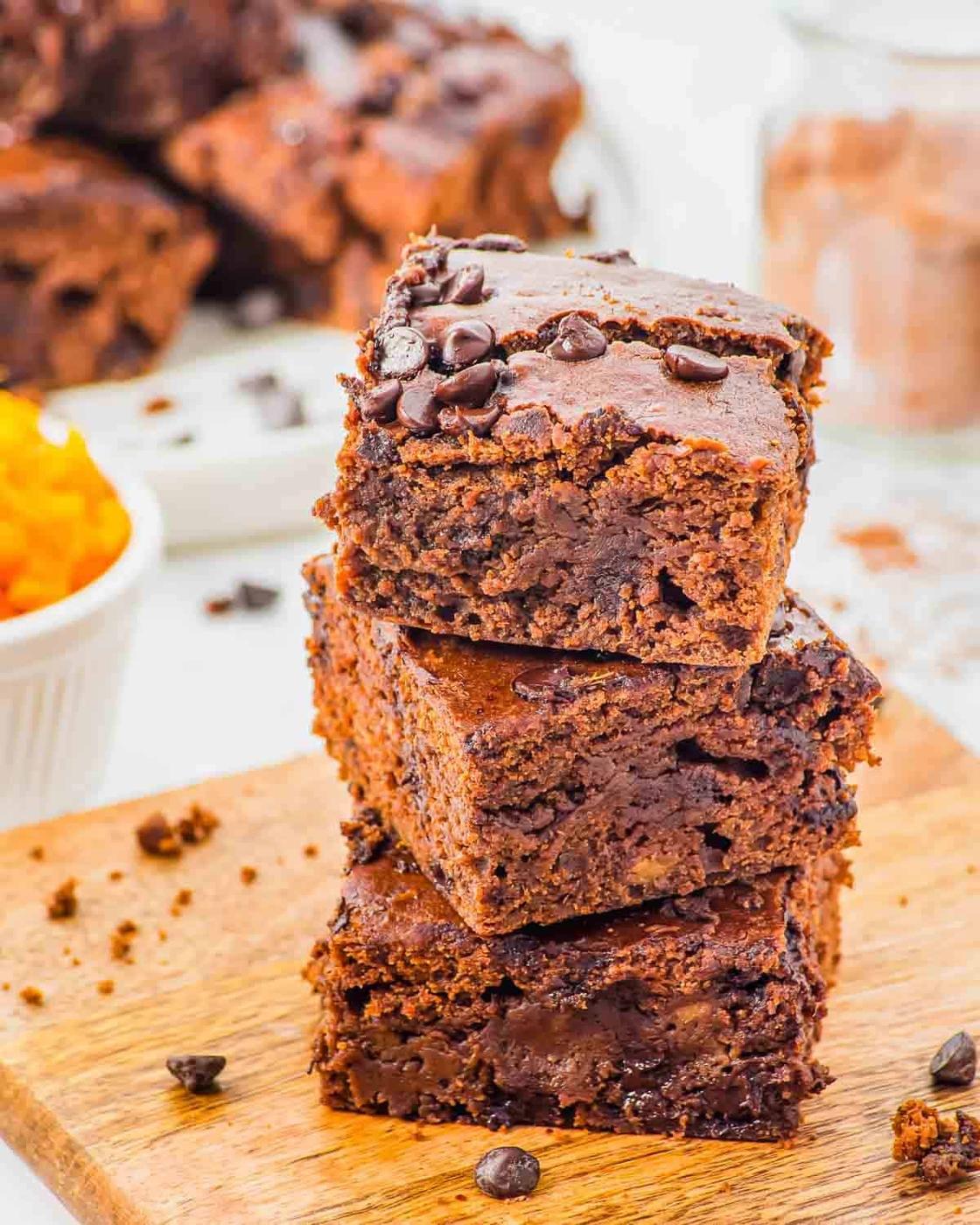Three sweet potato brownies stacked on a wooden board, topped with chocolate chips. The brownies look fudgy and rich, with more brownies and cocoa powder blurred in the background.