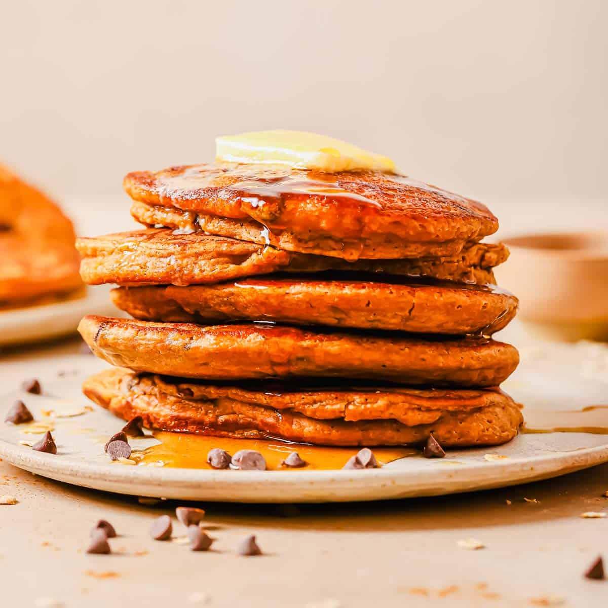 A stack of six pancakes topped with a pat of butter and drizzled with syrup, surrounded by chocolate chips on a beige plate.