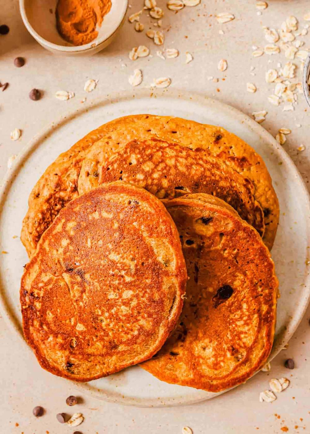 A plate of four golden-brown sweet potato chocolate chip pancakes with visible oats, placed on a light-colored surface scattered with oats and chocolate chips. A small bowl of cinnamon lies above the plate.