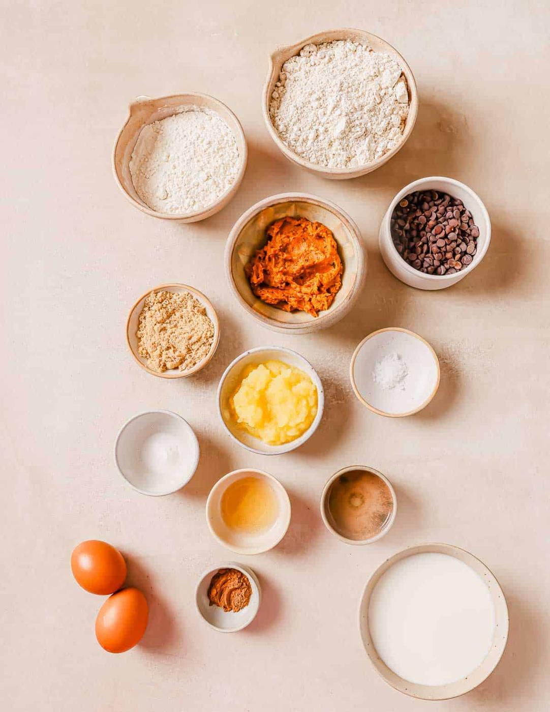Various ingredients in bowls and plates for baking, including flour, chocolate chips, brown sugar, pumpkin puree, eggs, butter, milk, spices, and baking powder arranged on a neutral surface.