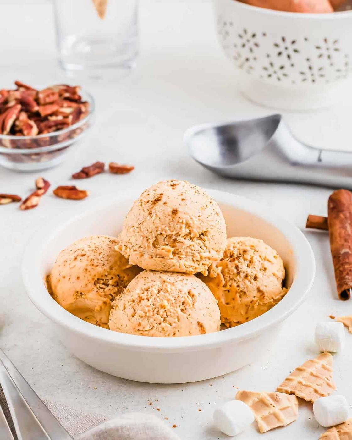A white bowl filled with four scoops of sweet potato ice cream, surrounded by an ice cream scoop, pecan pieces, and cinnamon sticks on a white surface.