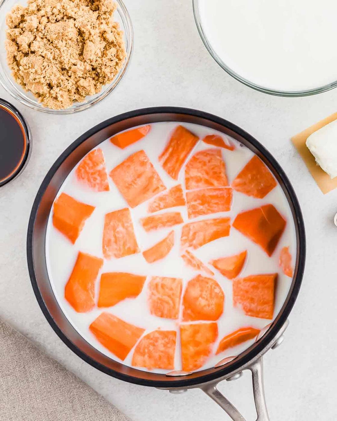A saucepan filled with sliced orange sweet potatoes and milk is surrounded by small bowls containing brown sugar, cream, and a rectangular block of cream cheese on a light surface.