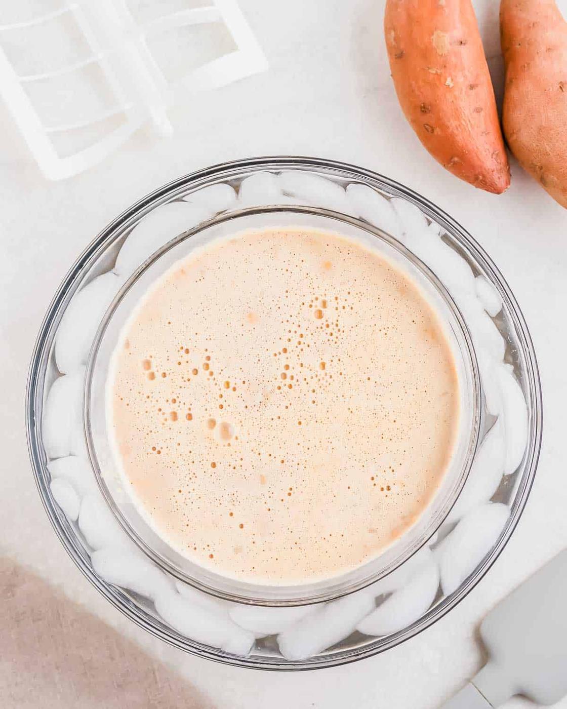 A bowl of ice cream base sits in a larger bowl filled with ice cubes. Two whole sweet potatoes are positioned nearby.